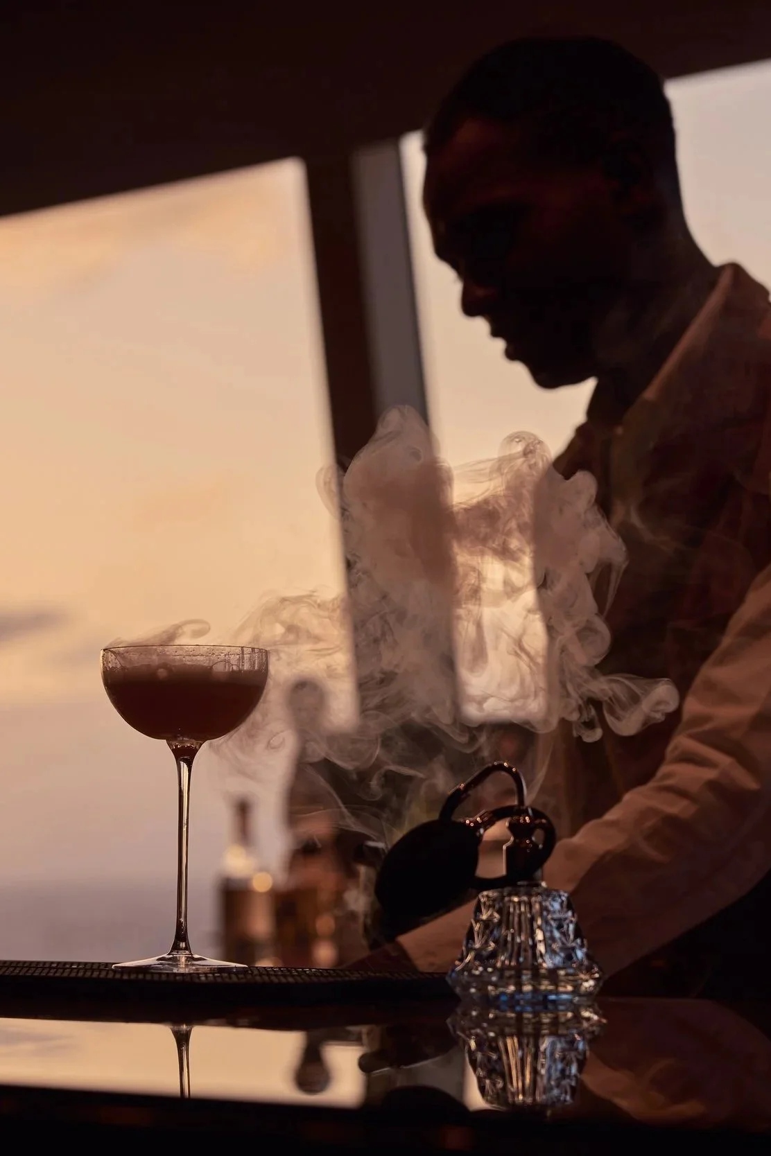 A silhouette of a bartender working with a smoking cocktail during sunset, with a glass of dark beverage on the bar.