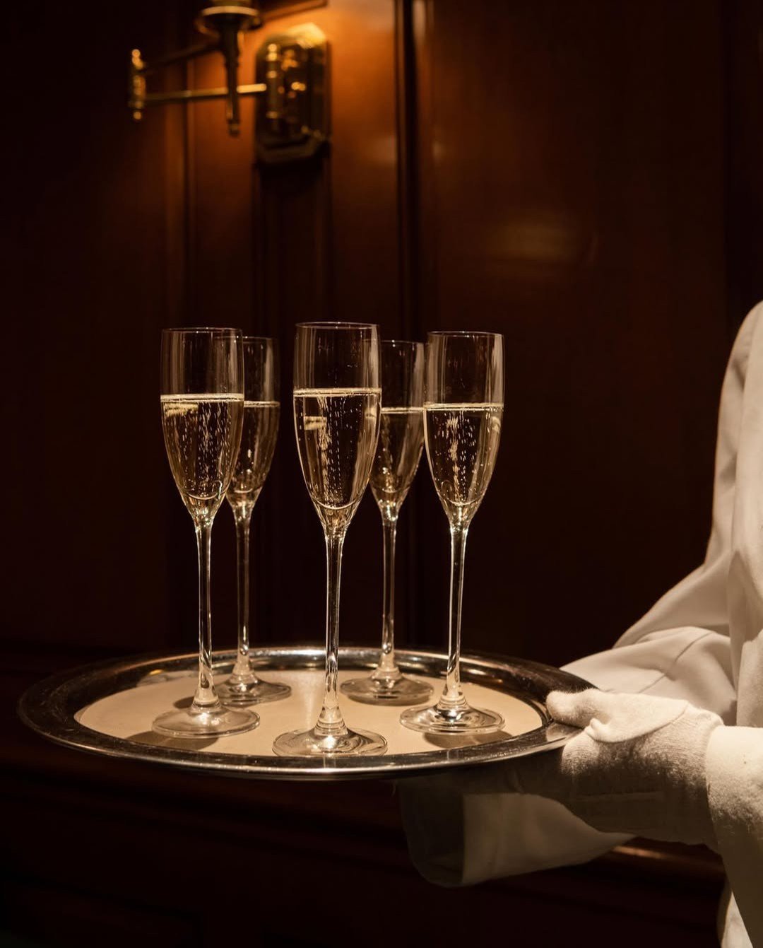 Five champagne flutes filled with bubbly champagne on a silver tray, being carried by a person wearing a white glove, against a warm, dark wooden background.