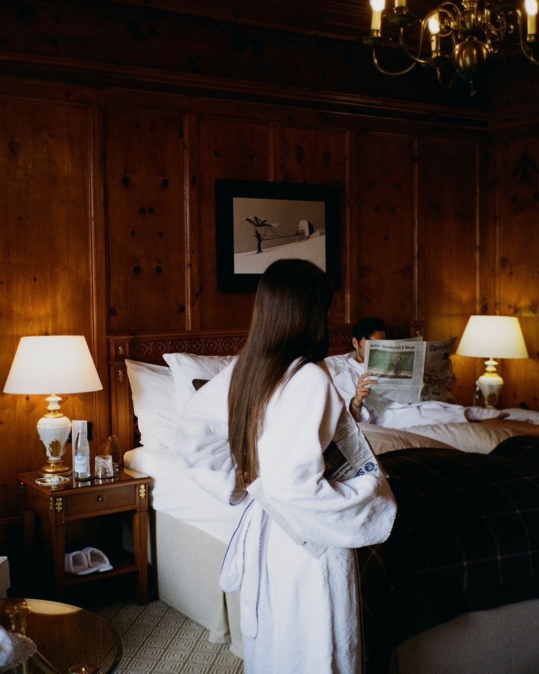 Two people in bathrobes in a warmly lit hotel room, one reading a newspaper and the other sitting on the bed, with wooden panel walls, bedside lamps, and a framed artwork overhead.
