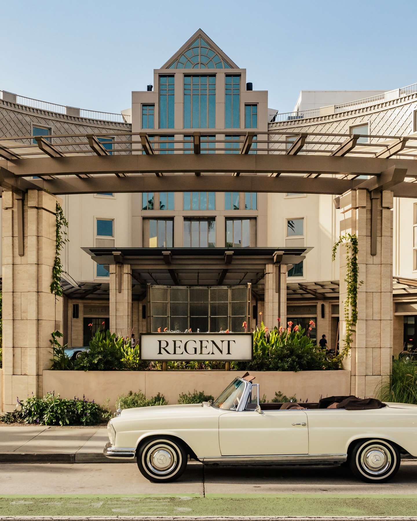 A vintage white convertible car parked in front of the Regent hotel with greenery and flowers, under a modern building with a sign that reads 'REGENT'.