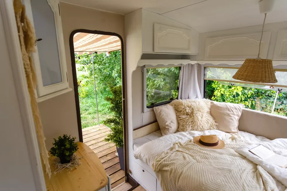 Cozy bed with white pillows and beige blanket inside a small camper or tiny house, with open door leading to a green outdoor garden area.
