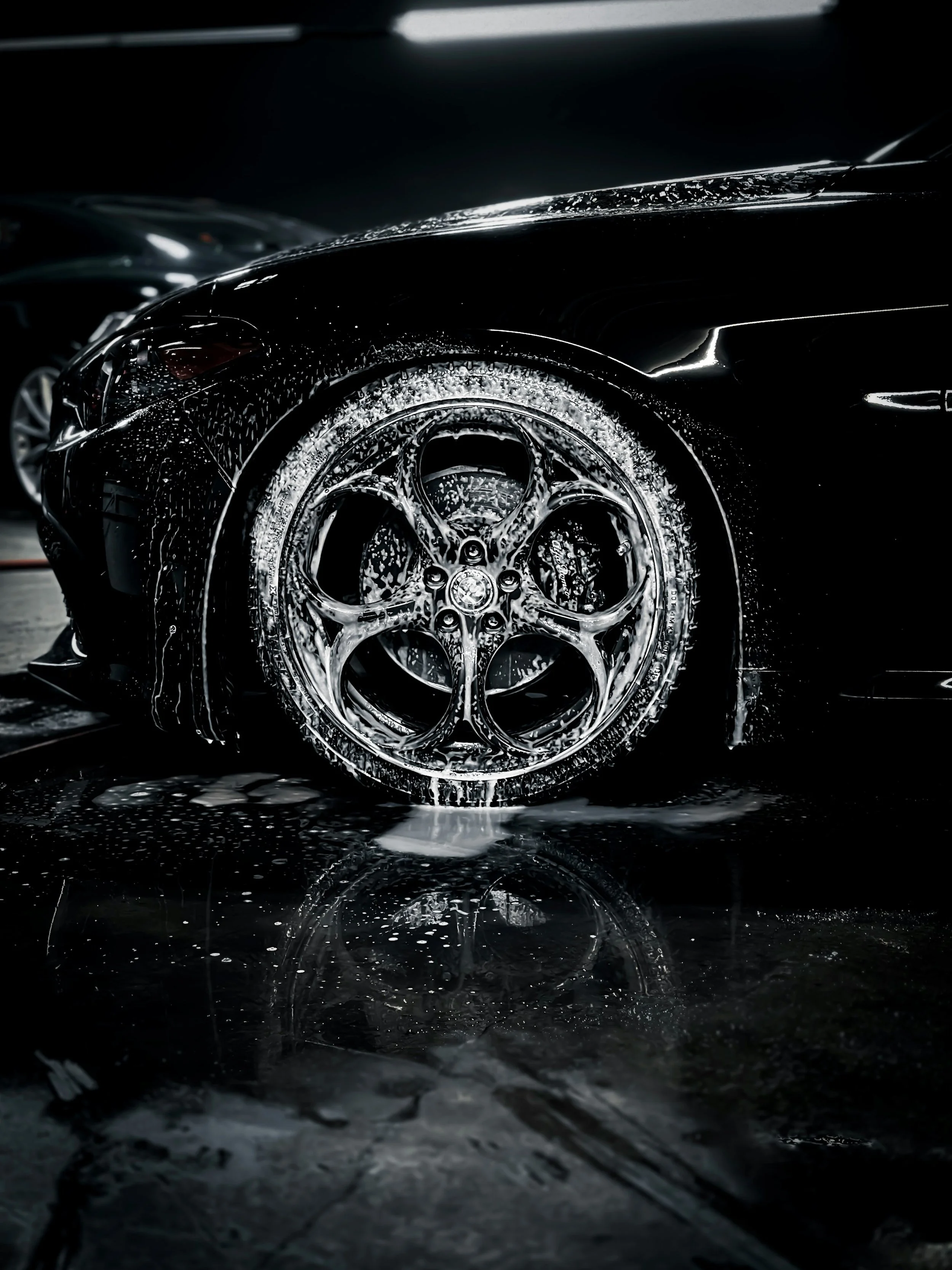 Close-up of a black luxury car being washed with soap, with water and soap suds on the car and reflections on the wet floor.