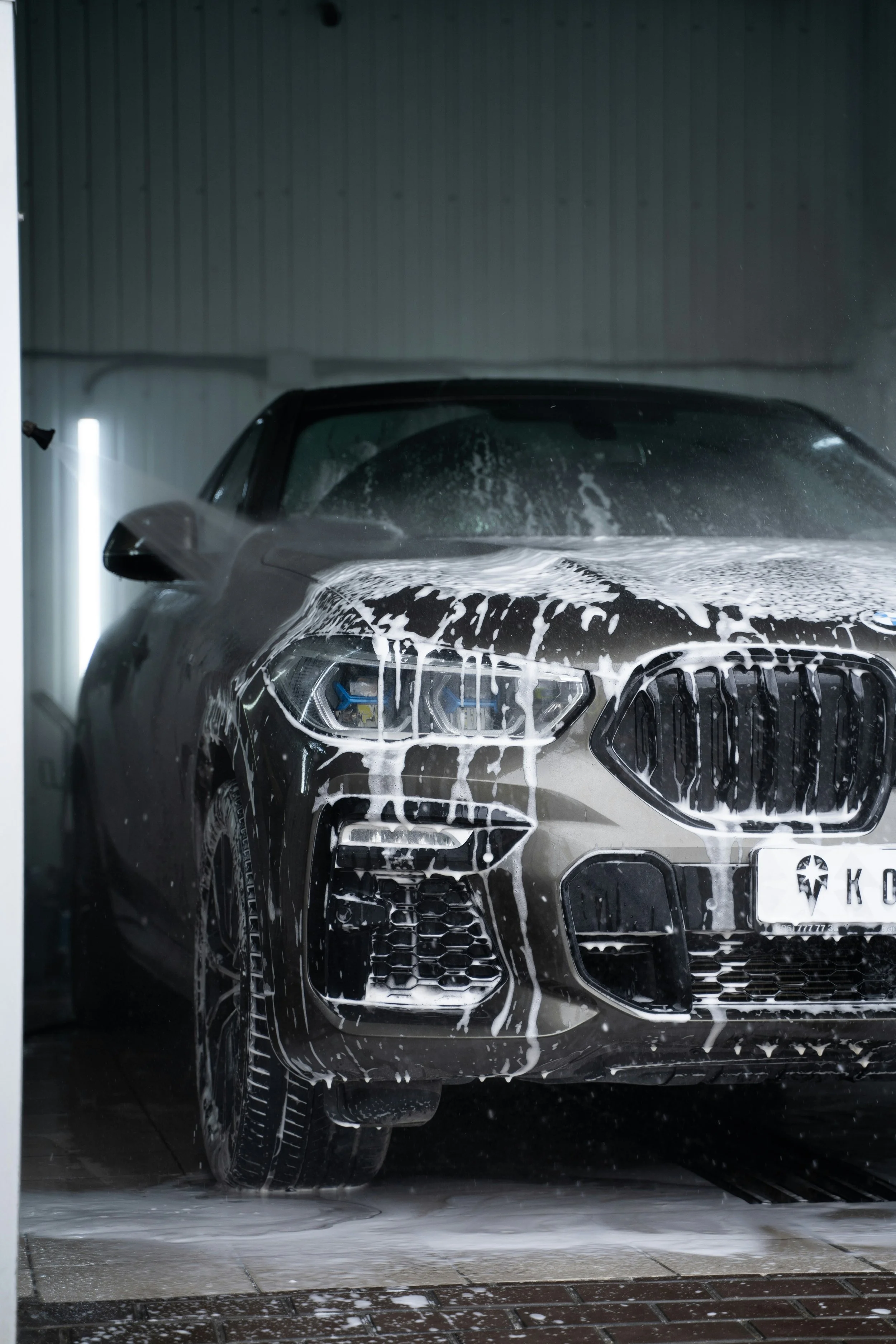 A silver luxury car, possibly a BMW, is in a car wash with soap and foam on its front and windshield.