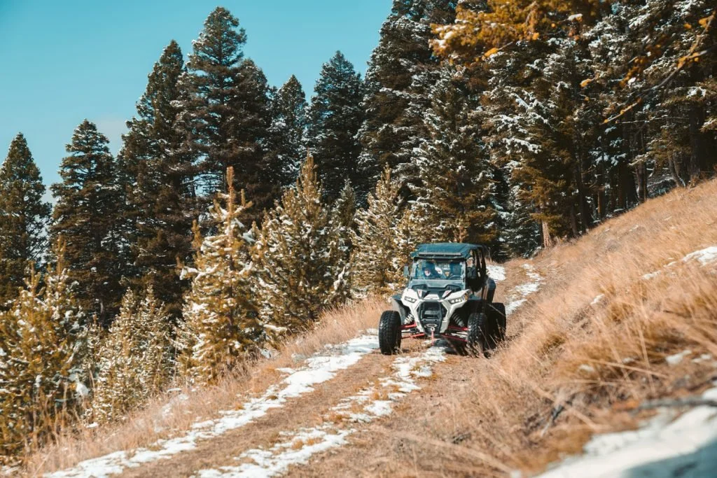 An off-road vehicle driving on a dirt trail through a snowy pine forest on a clear day.