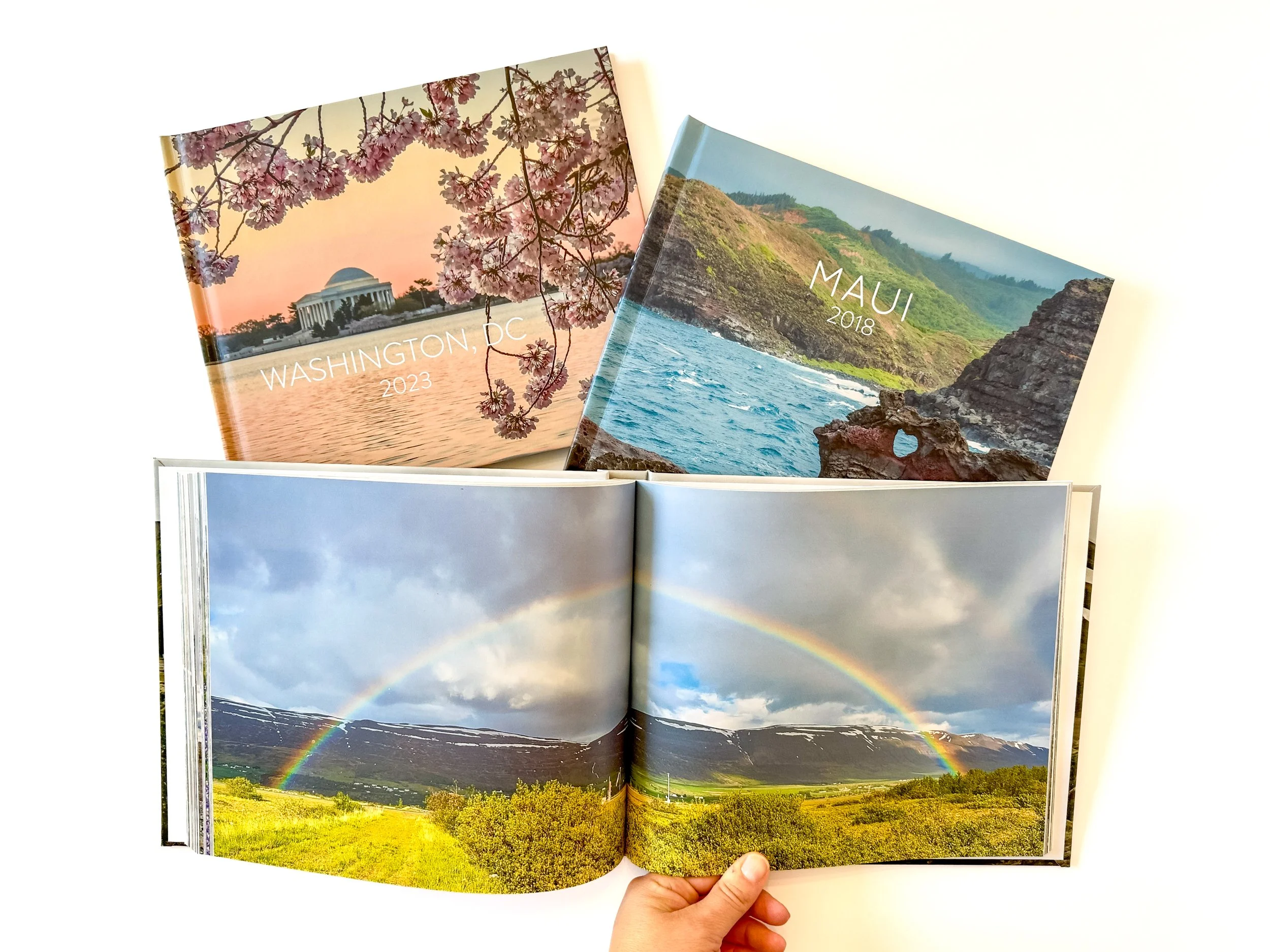 Three travel photo books with scenic landscape covers, one open showing a rainbow over green fields and mountains.