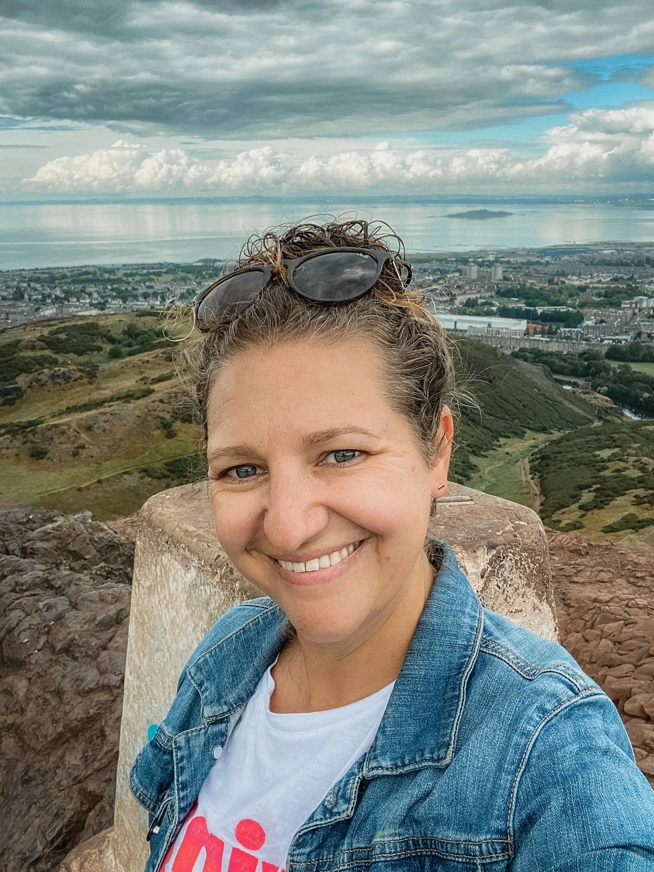 A woman taking a selfie on a hilltop with a scenic view of a city, green hills, and a body of water in the background. She is smiling, wearing a denim jacket, and has sunglasses on her head.
