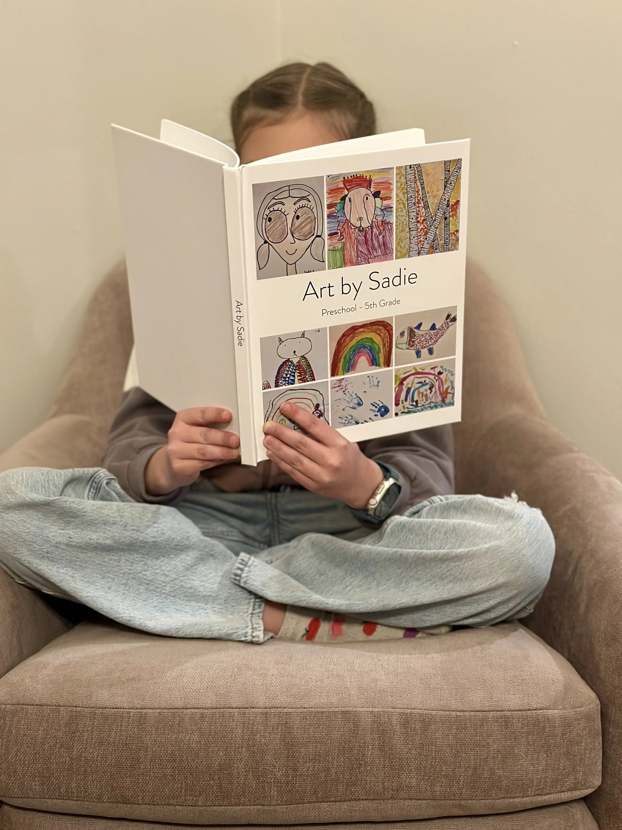 A child sitting cross-legged on a beige armchair, holding and reading a book titled 'Art by Sadie,' with colorful illustrations on the cover.