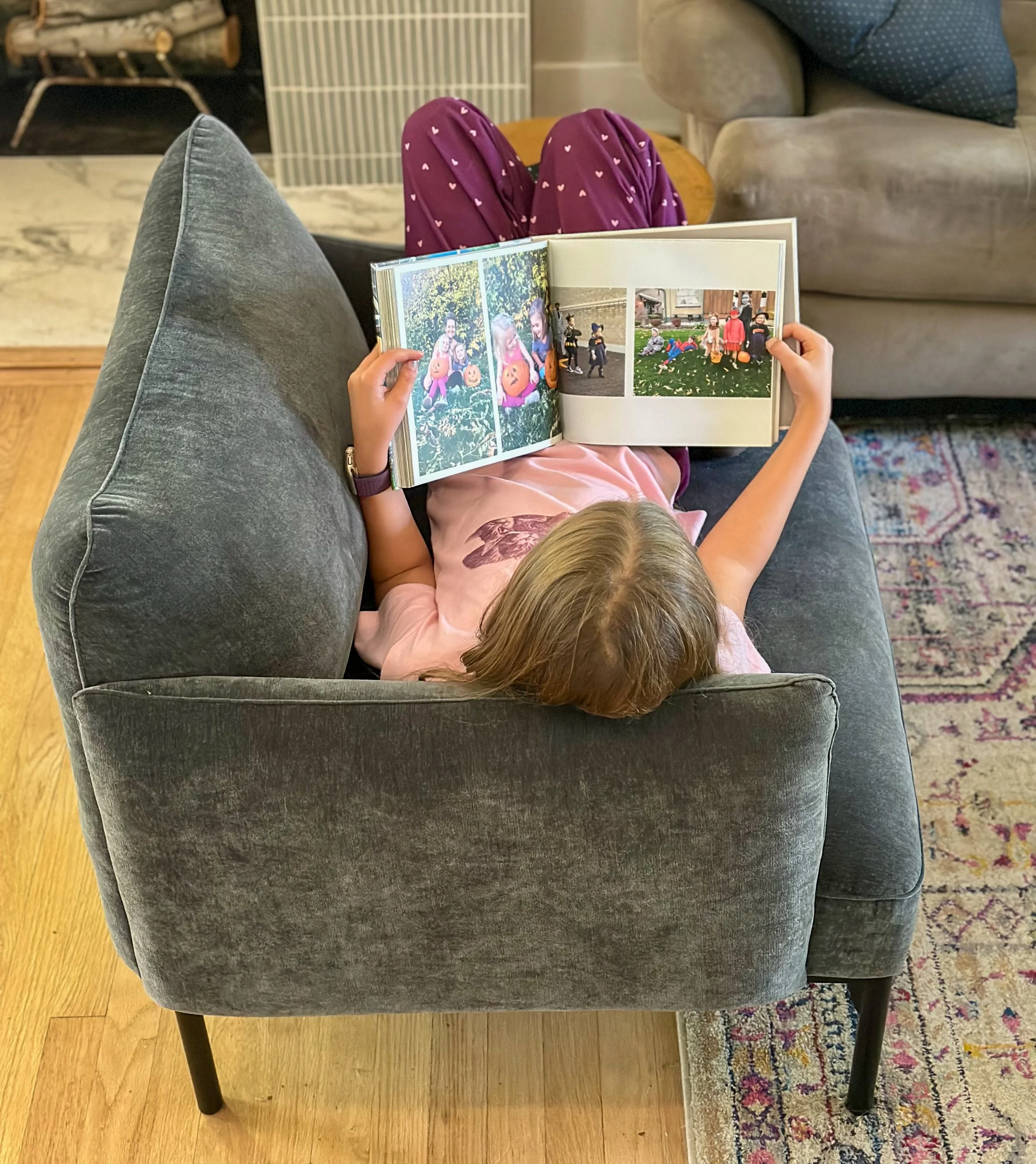 A person lying on a gray couch, reading a photo album. The person is wearing a pink shirt with a floral design and has light brown hair. The photo album has pictures of children playing outside.