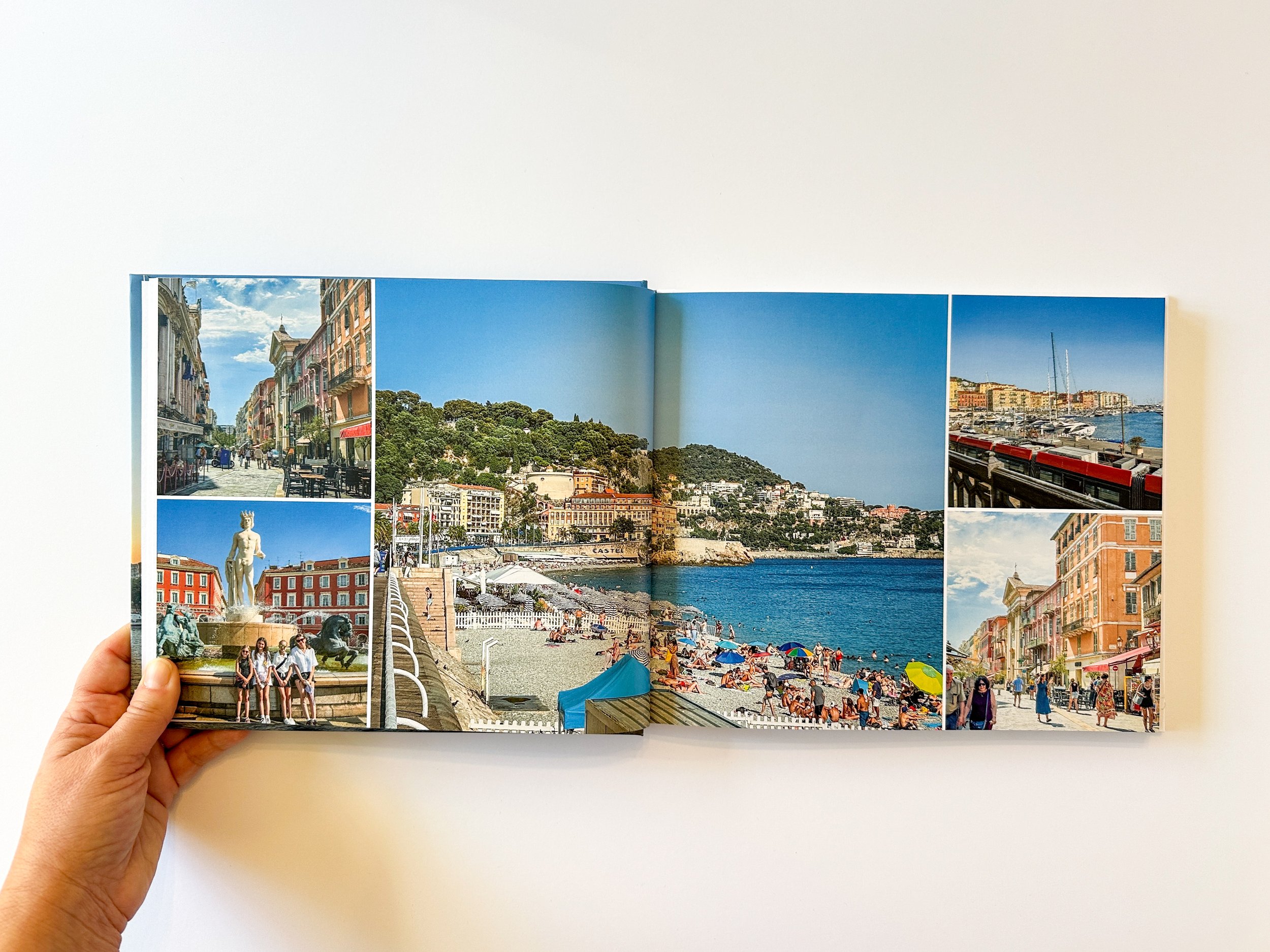Open photo book with Mediterranean coastal scenes including a street with colorful buildings, a hill with greenery, a beach with umbrellas and people, a marina with boats, and statues in a plaza, shot in bright daylight.