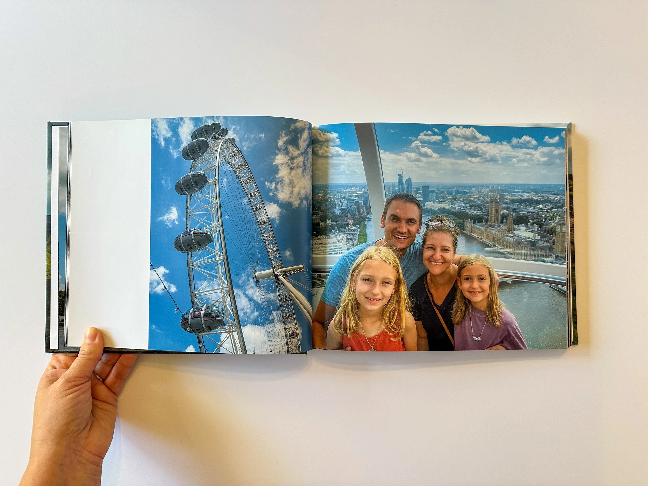 A person holding open a photo album showing a photo of a The London Eye on the left page and a family of four taking a selfie with the London skyline in the background on the right page.