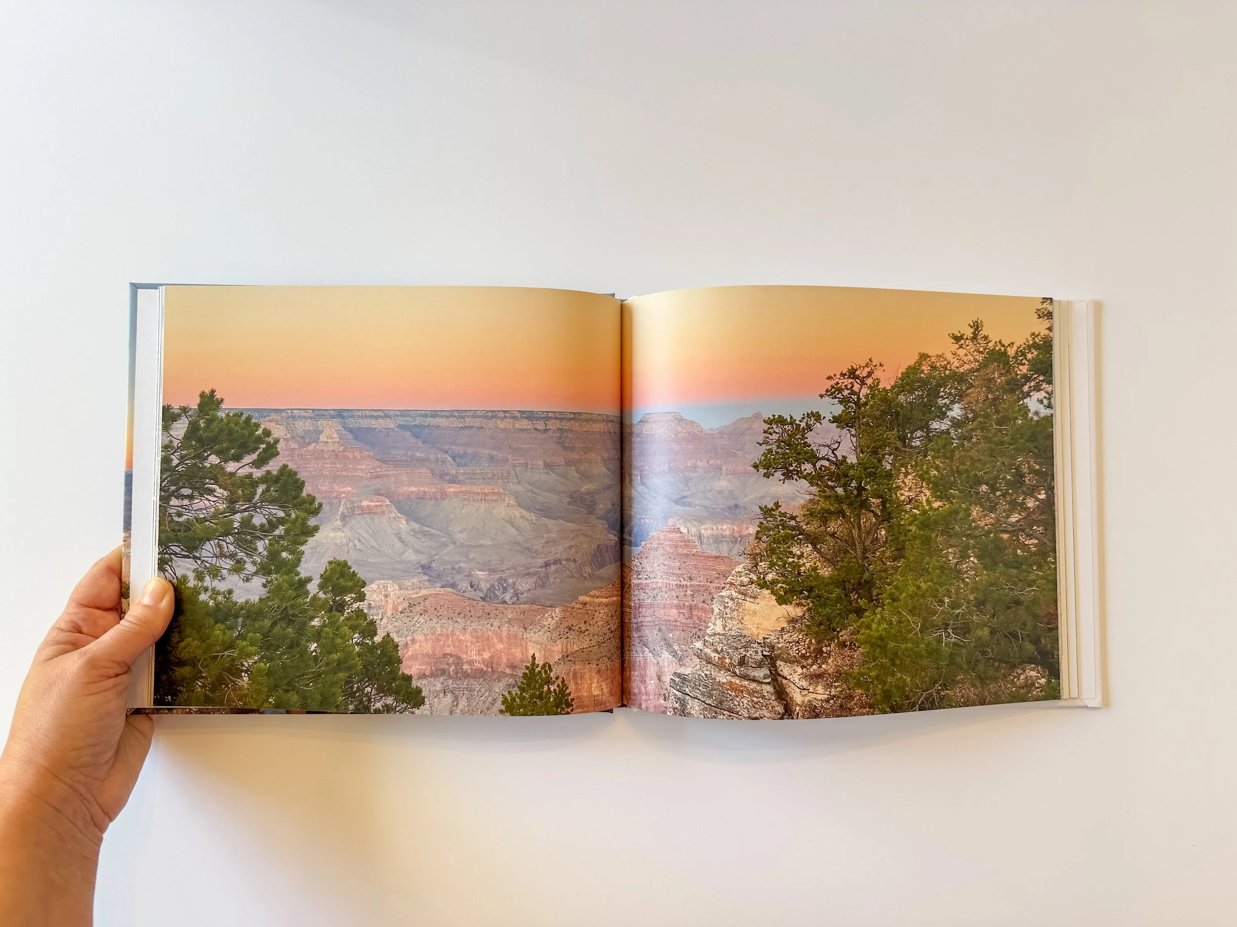 Open photo book showing a scenic view of the Grand Canyon at sunset, with trees in the foreground and colorful sky.