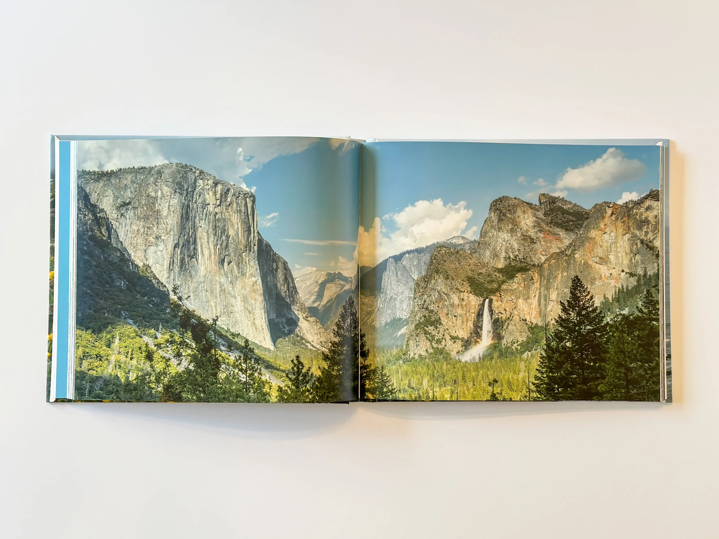 Open photo book showing a mountain landscape in Yosemite with tall cliffs, green trees in the foreground, a waterfall, under a partly cloudy sky.