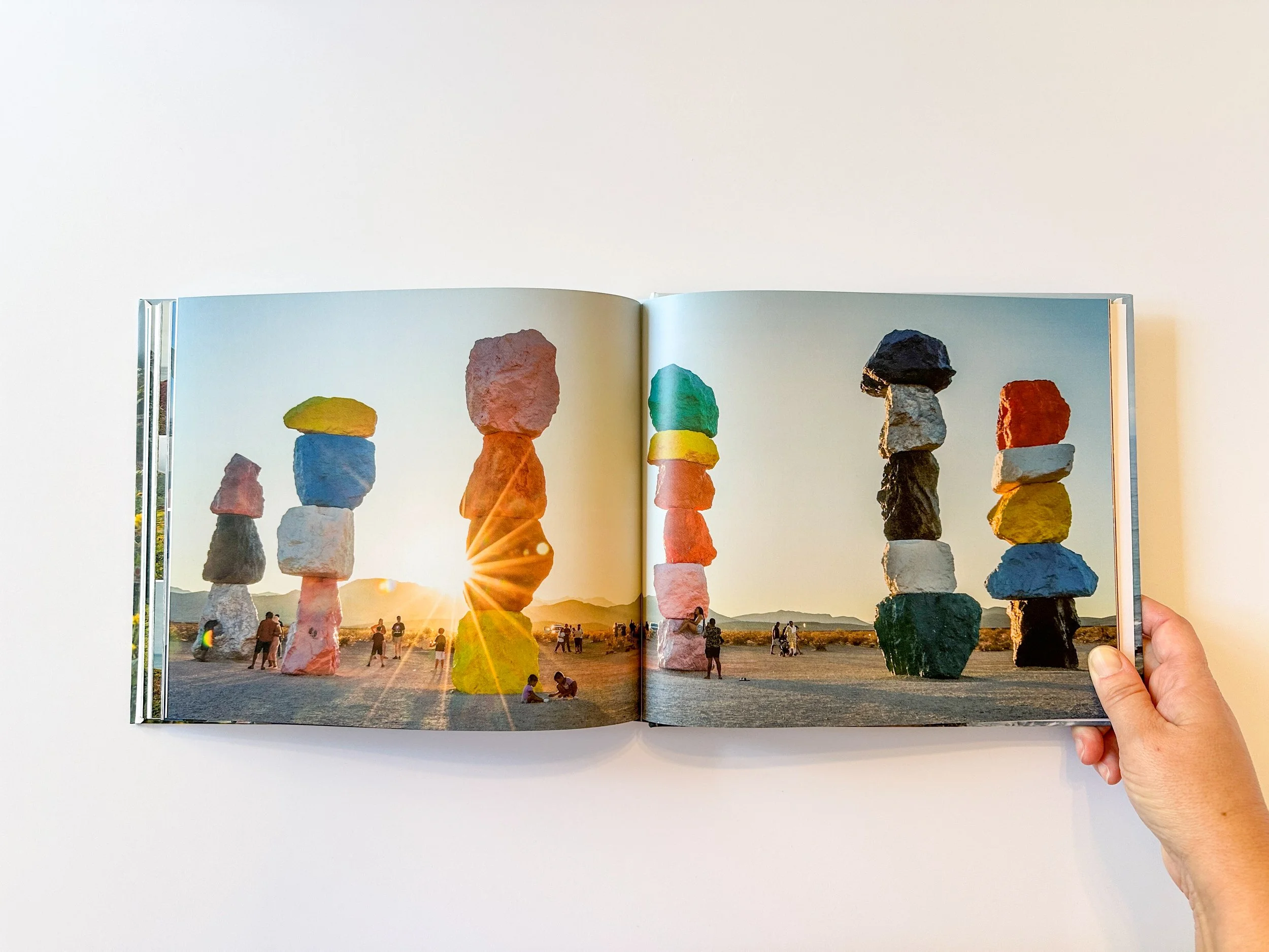 A person holding an open book displaying a photo of large stacked rocks or stone sculptures at sunset, with people walking and sitting on the ground and distant mountains in the background.