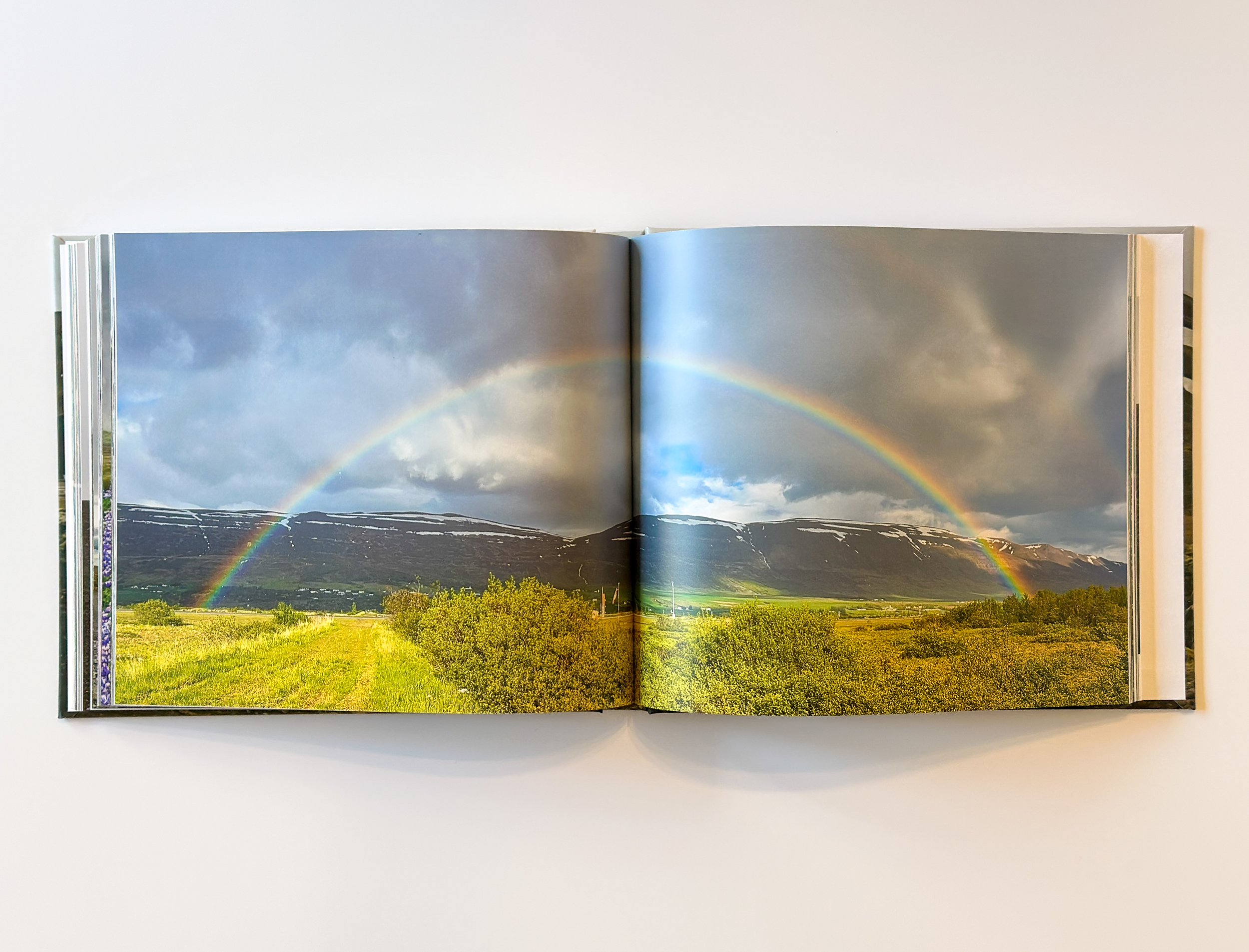 Open photo book displaying a landscape with double rainbow over green fields and mountains under a cloudy sky.