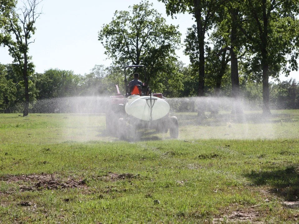 A person riding a tractor with a sprayer attachment across a grassy field, spraying herbicides and pesticides on the grass.