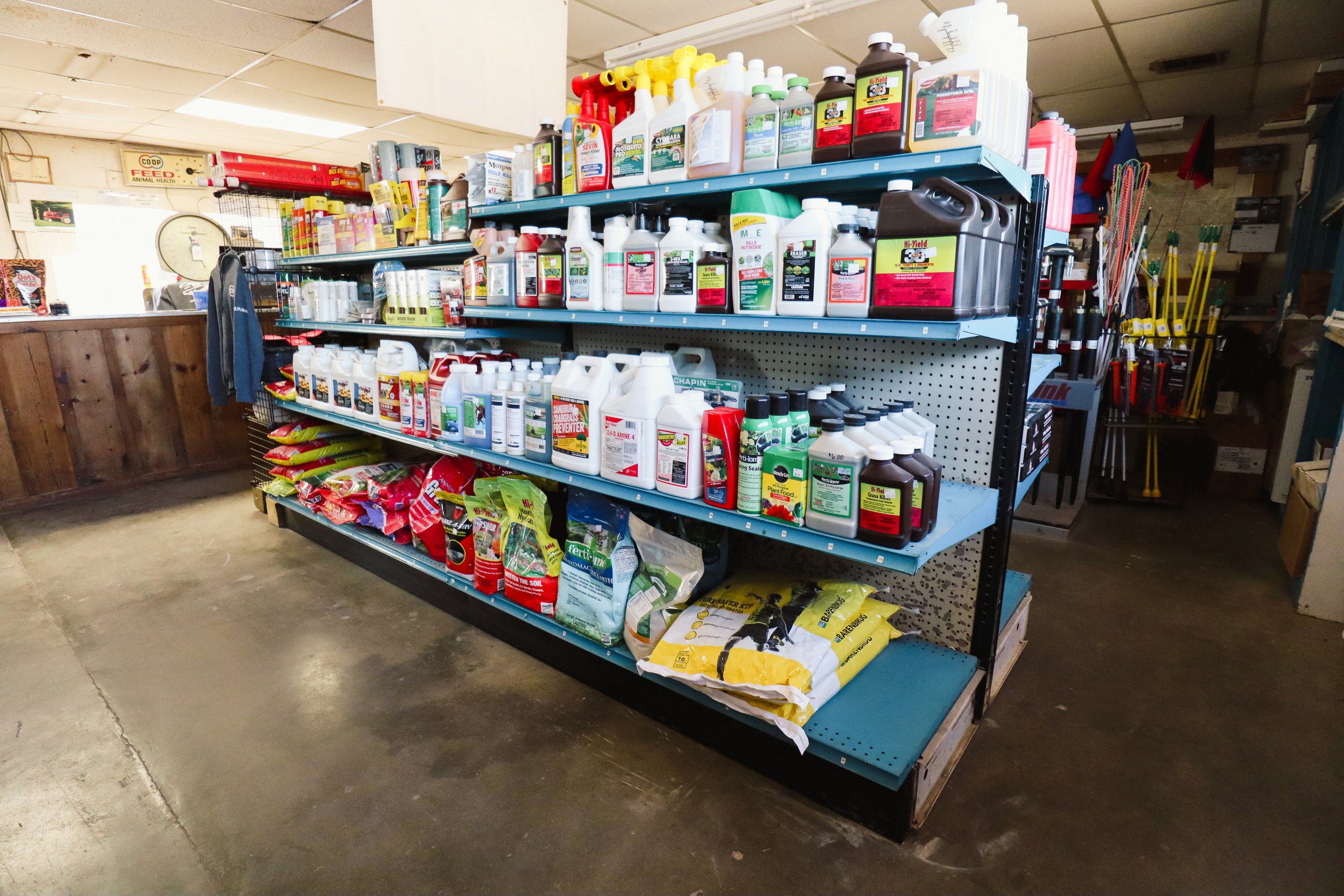 A store shelf stocked with various bottles of garden chemicals and fertilizers, with gardening tools and supplies in the background.