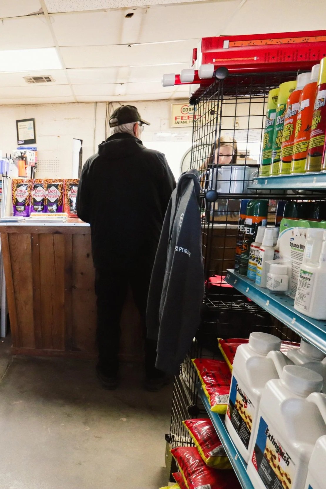 An elderly man with white hair, wearing a black jacket and cap, stands at a store checkout counter. There are shelves with pet supplies and food on his right, while a cashier with long hair, wearing glasses, assists him behind the counter. The store