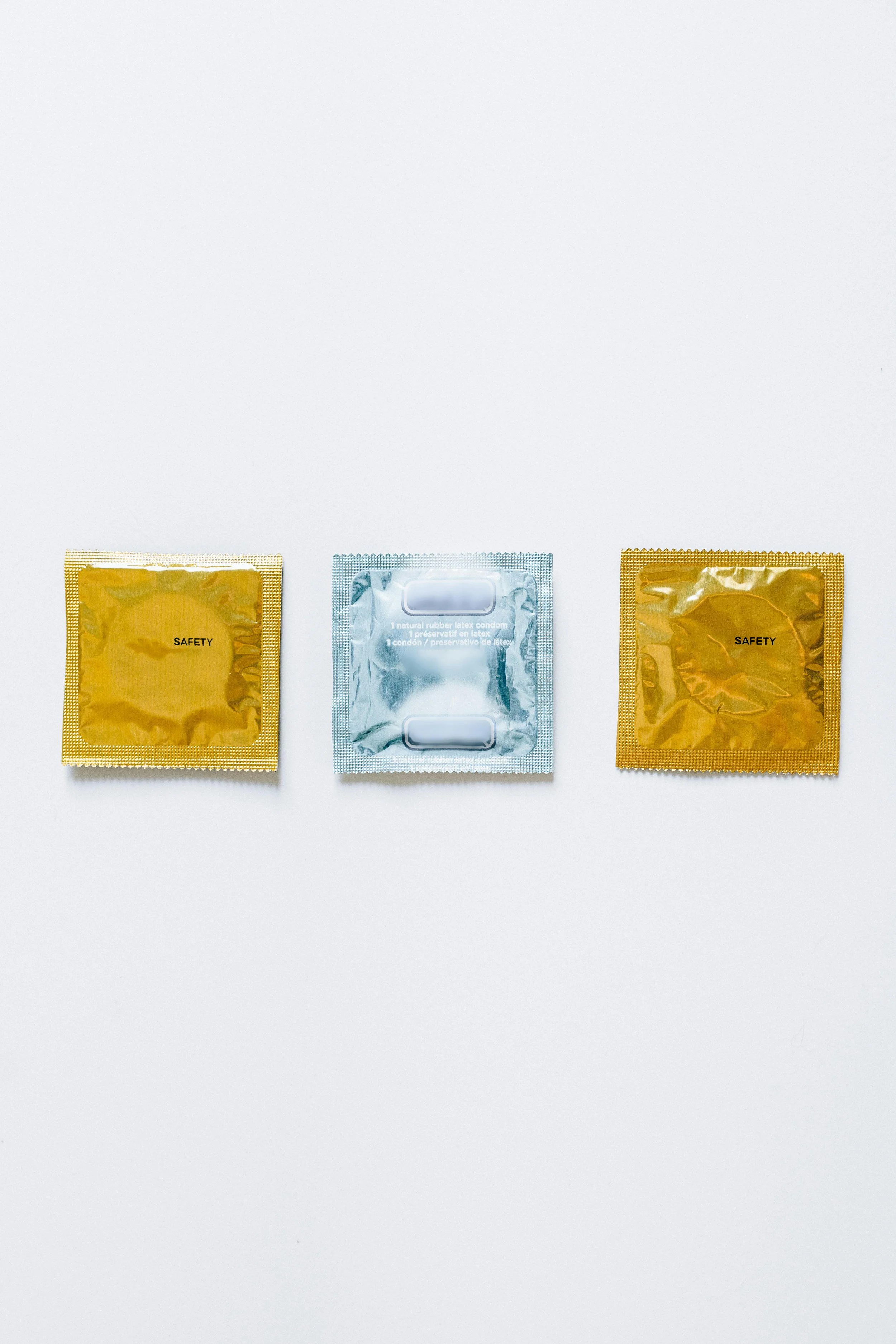 Drei Kondome in Verpackungen auf weißem Hintergrund, zwei goldene mit der Aufschrift ‚Safety‘, eins silberne mit Verpackungshinweisen.