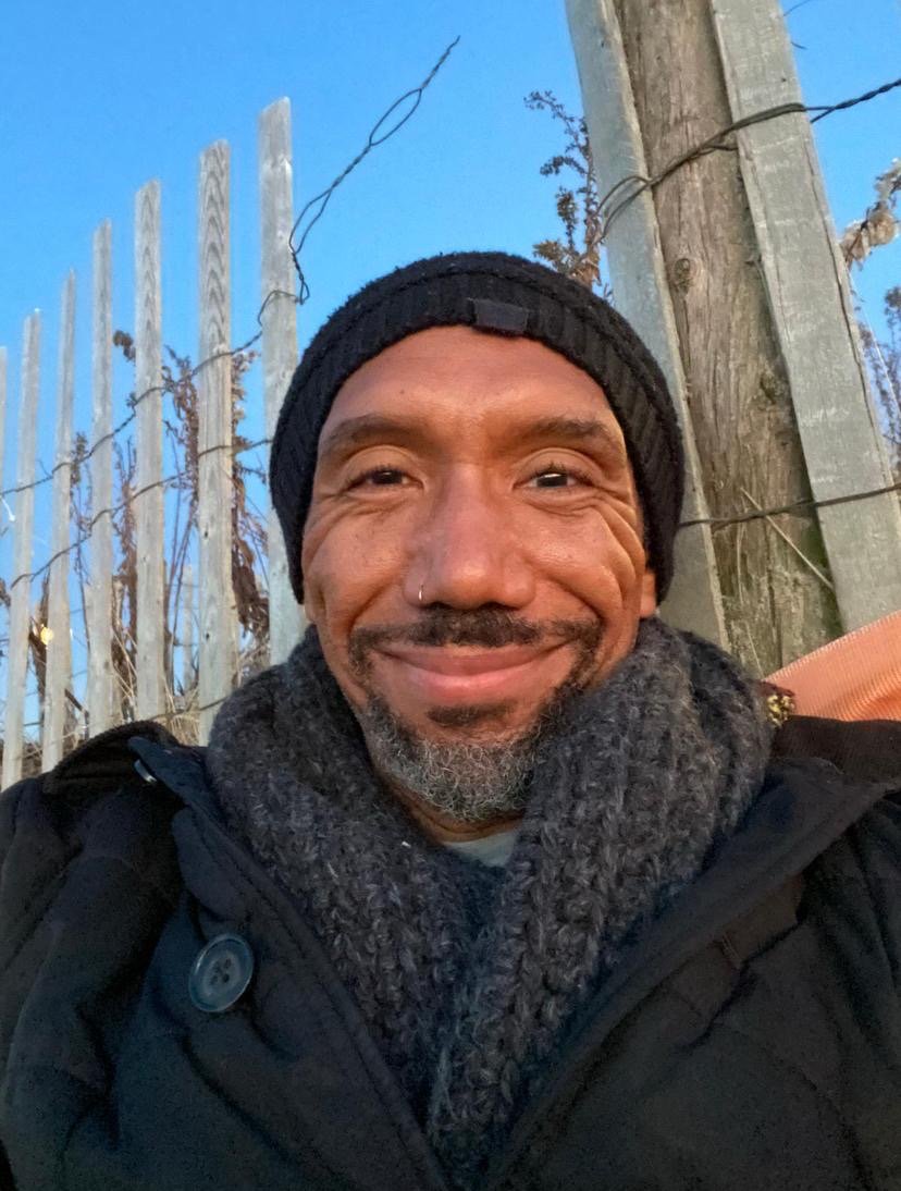 Close-up of a smiling man with a dark beard and mustache, wearing a black knit hat and a dark coat, standing outdoors in front of a wooden fence with barbed wire and some trees. Clear blue sky in the background.