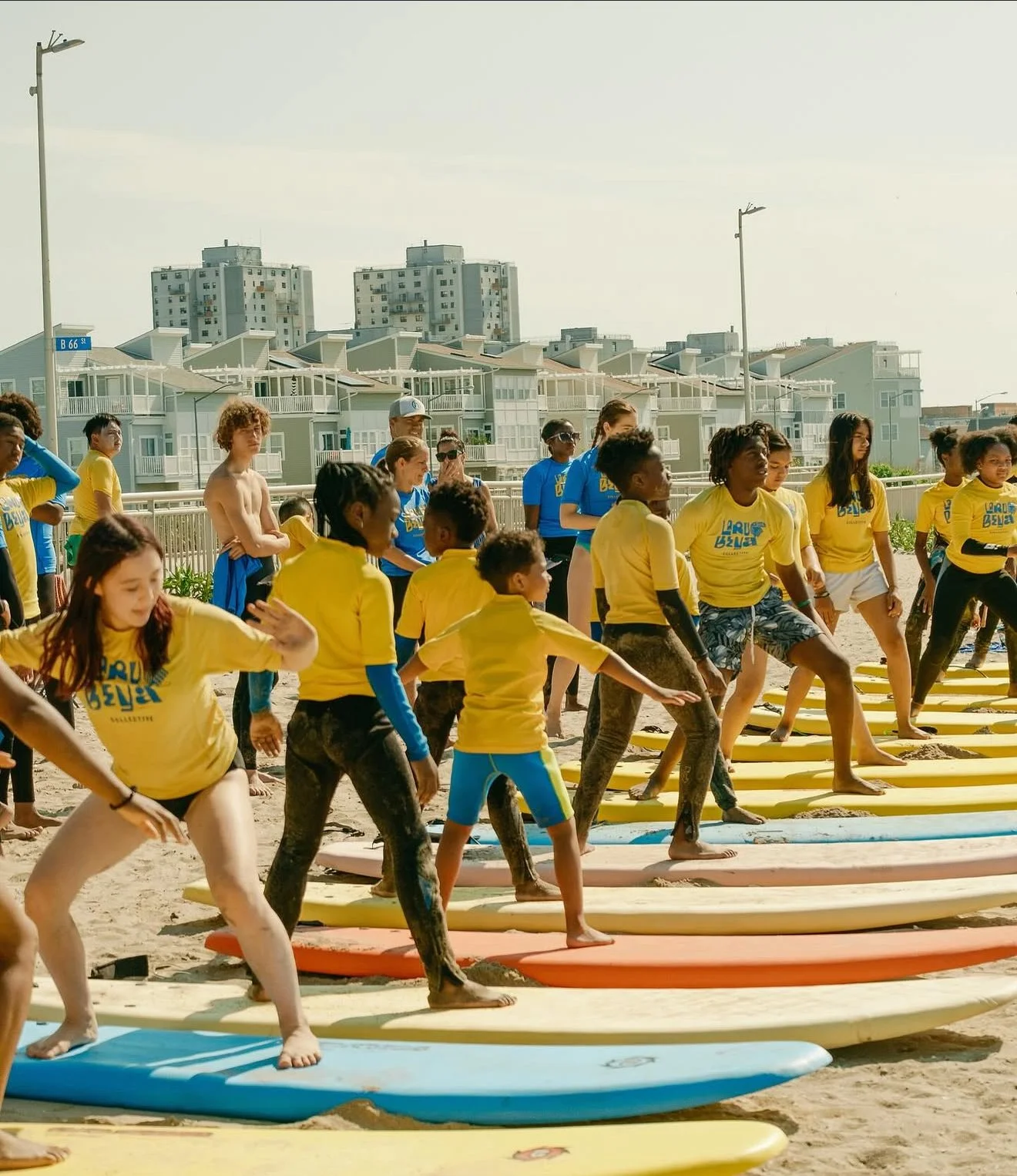 Group of children and teenagers participating in a surfing class on a sandy beach, some wearing yellow shirts, with surfboards laid out on the sand in front of them and residential buildings in the background.