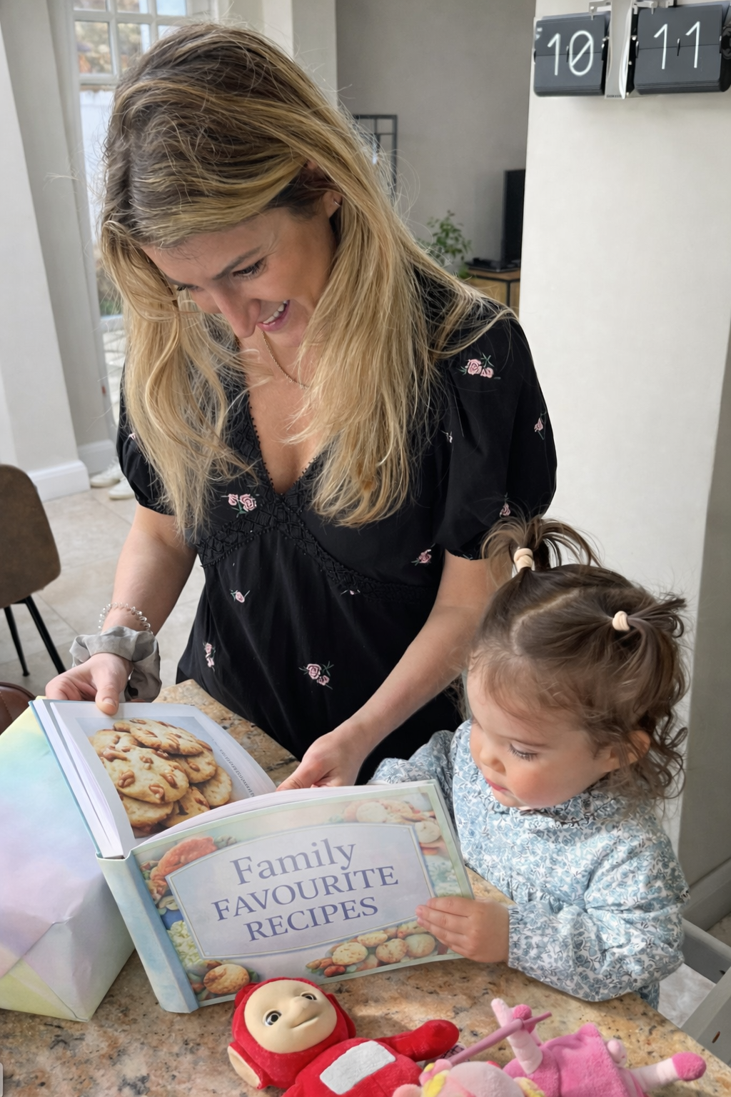 A woman with blonde hair and a young girl with pigtails looking at a cookbook titled 'Family Favourite Recipes' on a kitchen countertop. The woman is pointing to a page with pictures of cookies, and there are toys on the counter, including a plush doll and a pink unicorn.