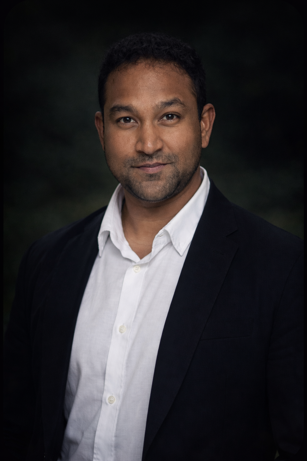 Headshot of a man with short dark hair, wearing a black suit jacket and white shirt, against a dark background.