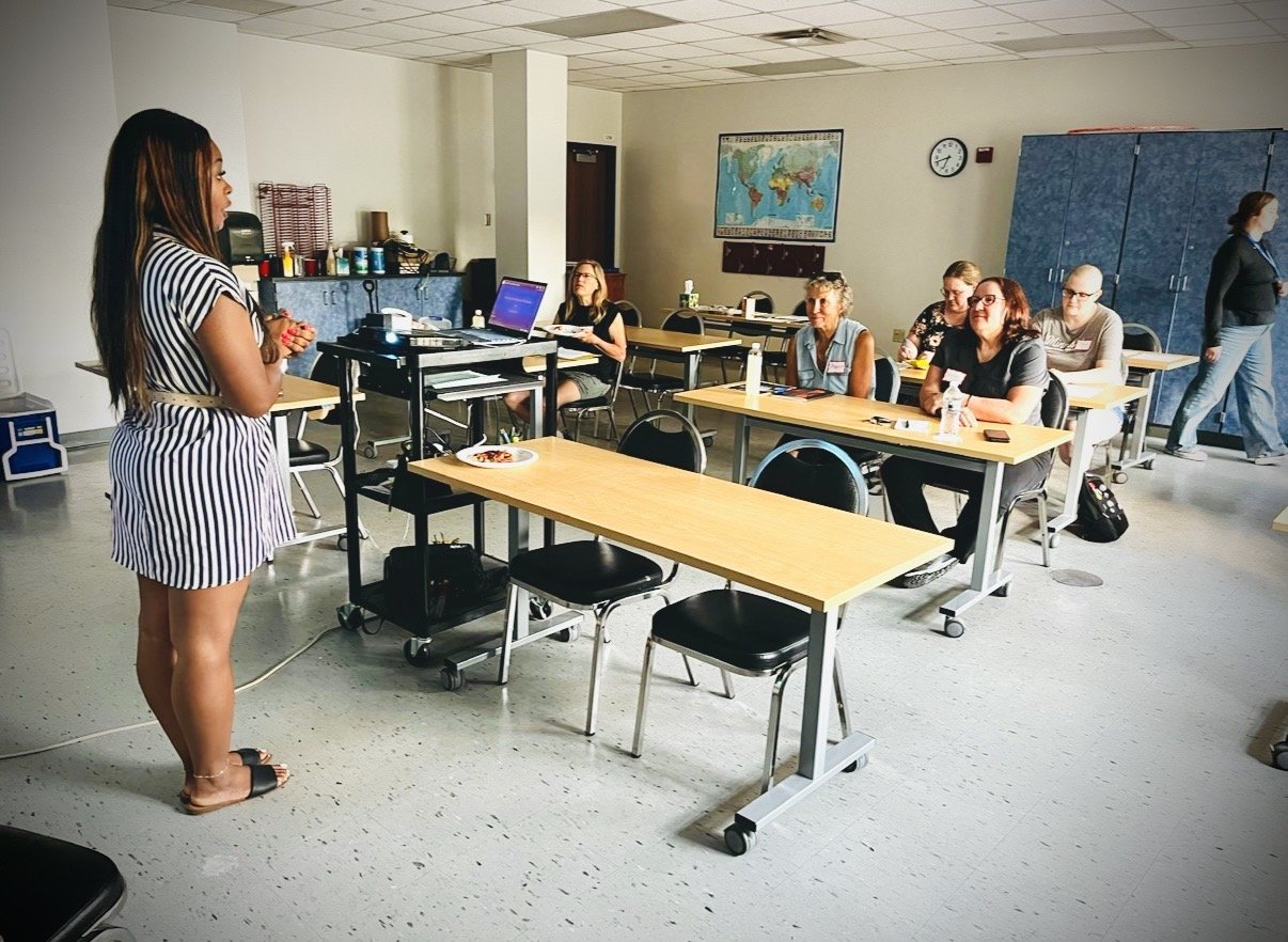 A woman standing at the front of a classroom giving a presentation to a seated audience of women in a conference room with world map on the wall.