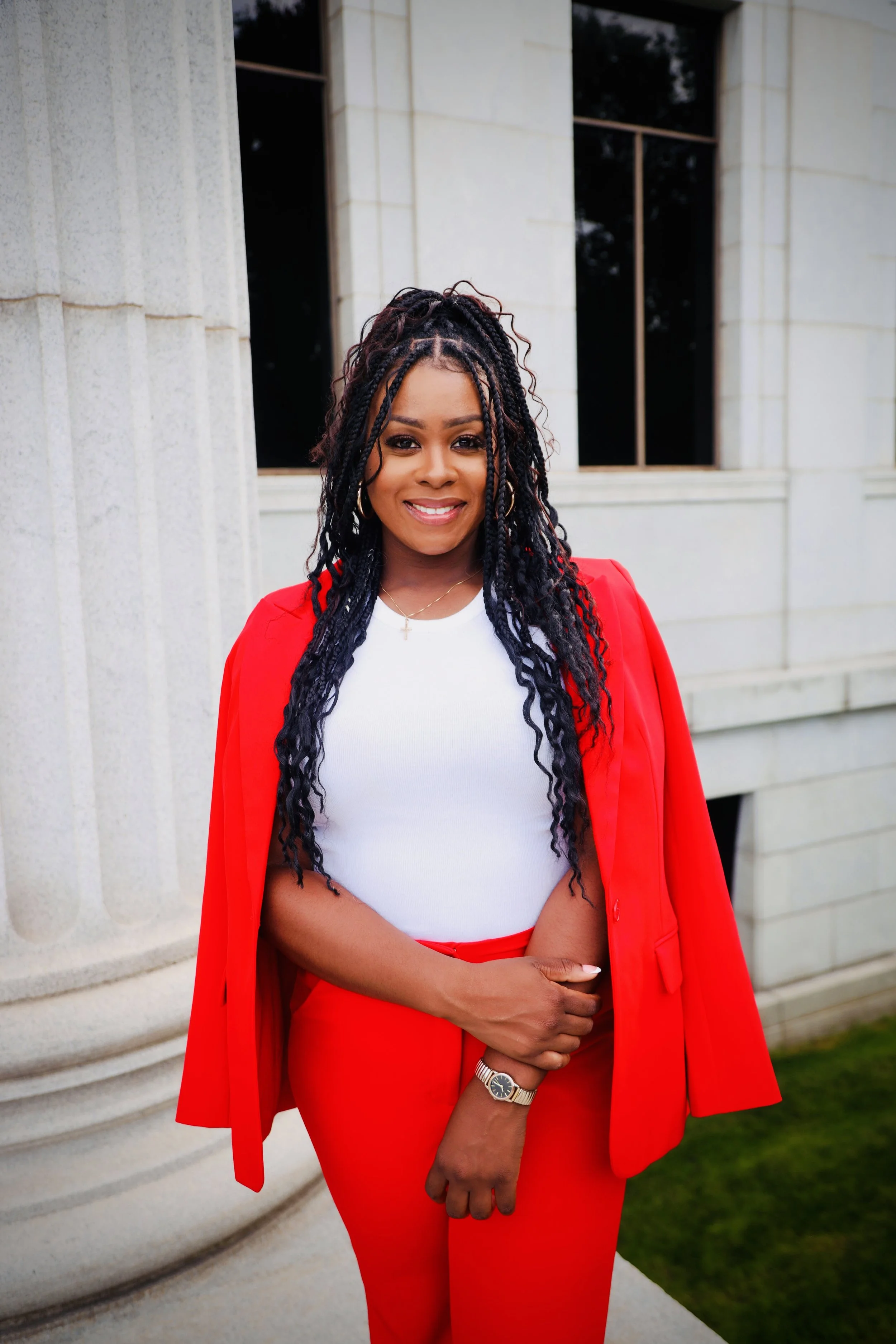 A smiling woman with natural curly hair styled in twists, wearing a bright red blazer and matching red pants, a white top, a wristwatch, and a delicate necklace, standing outdoors in front of a building with light-colored stone walls and large windows.