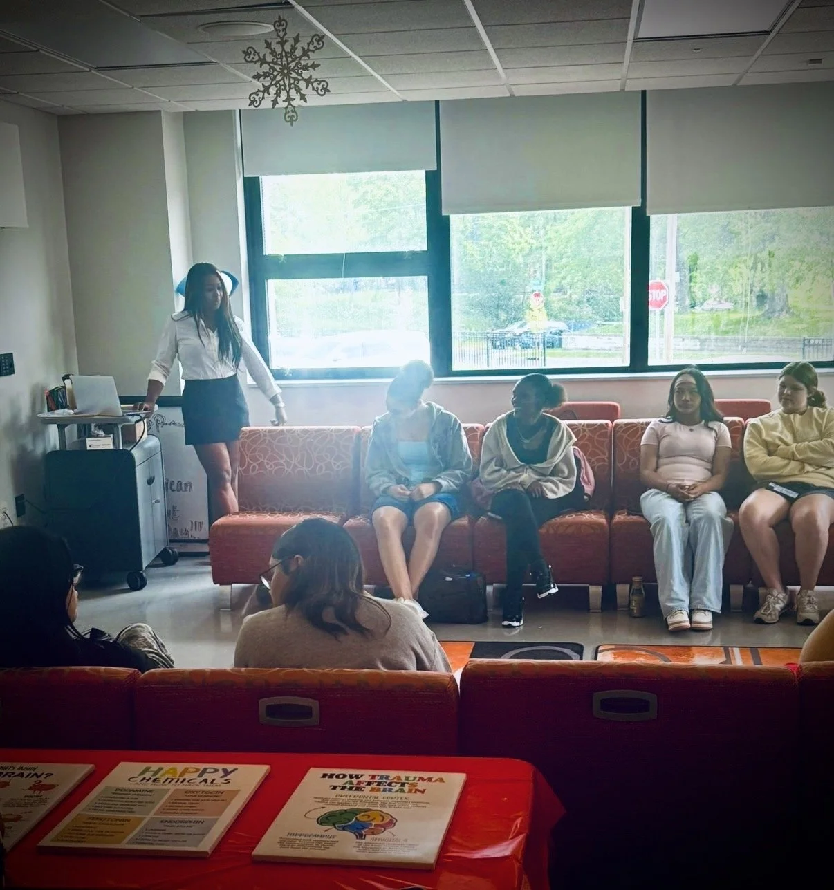 Group of five women sitting on a red-orange couch in a room with large windows; one woman standing near a small table and a laptop. The window shows trees and a stop sign outside.