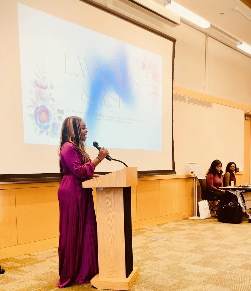A woman in a long purple dress is speaking at a podium with a microphone during a conference or event. Behind her, there is a large screen displaying the title 'Empower & Rise' along with some floral graphics. Two women are seated at a table to the right, facing the audience.