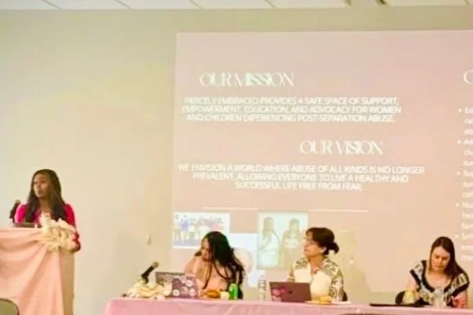 A woman standing at a pink podium on the left, three women sitting at a pink table on the right, a large screen behind them displaying text about empowerment, and two images of women on the screen.