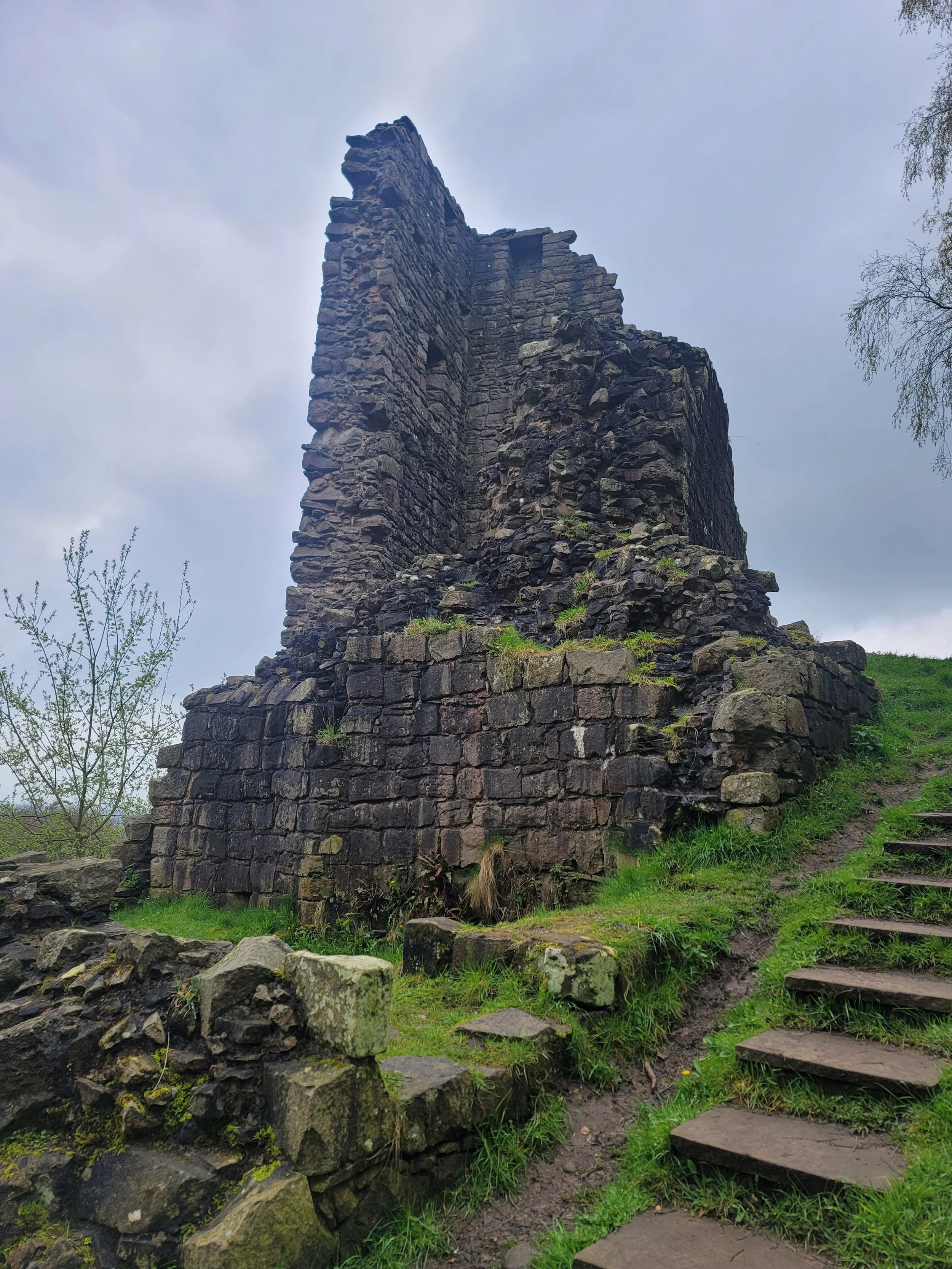 Ancient stone ruin on a grassy hill with stone steps and trees, under a cloudy sky.