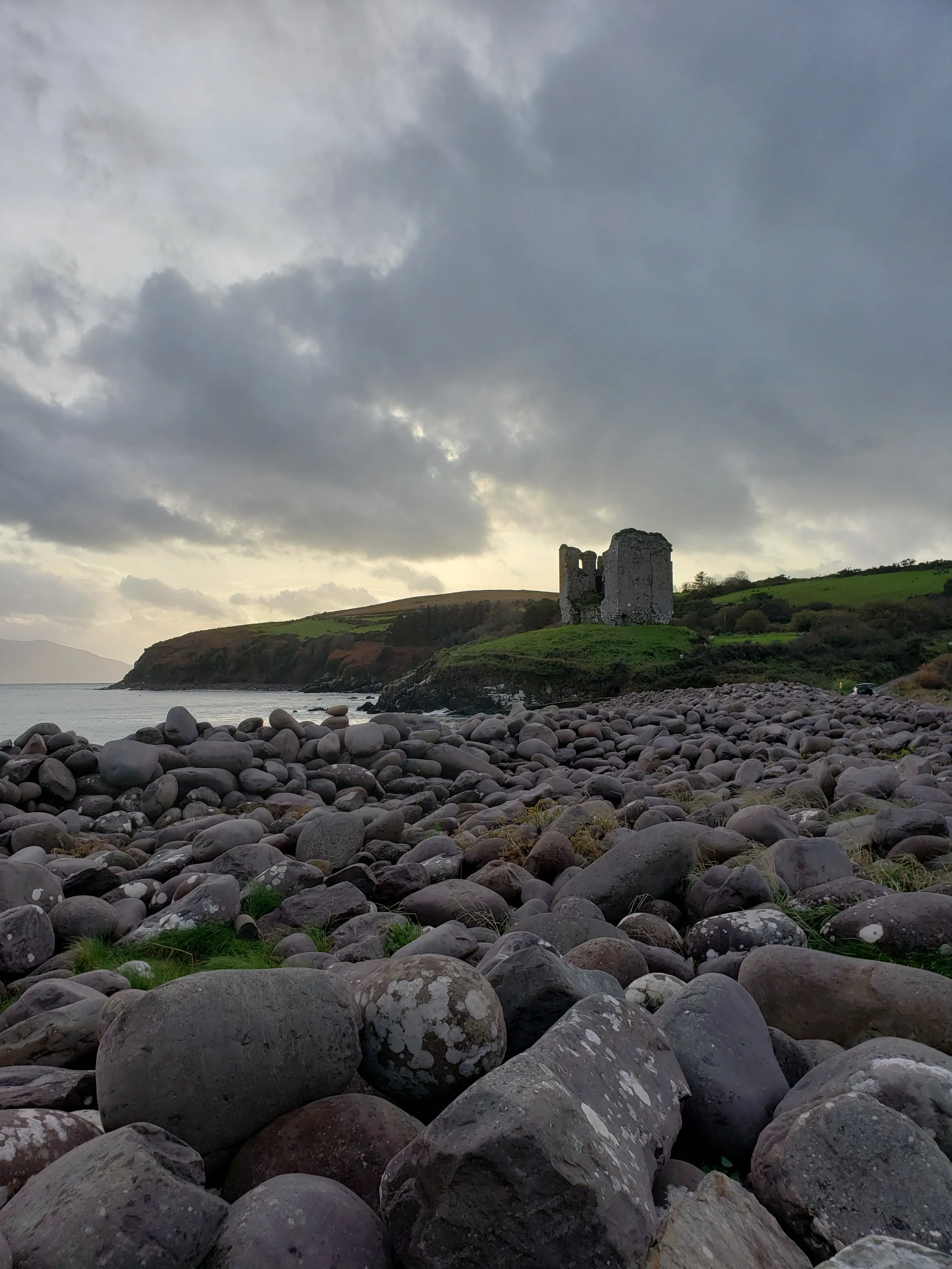 Rocky shoreline with a ruin on a green hillside under a cloudy sky.