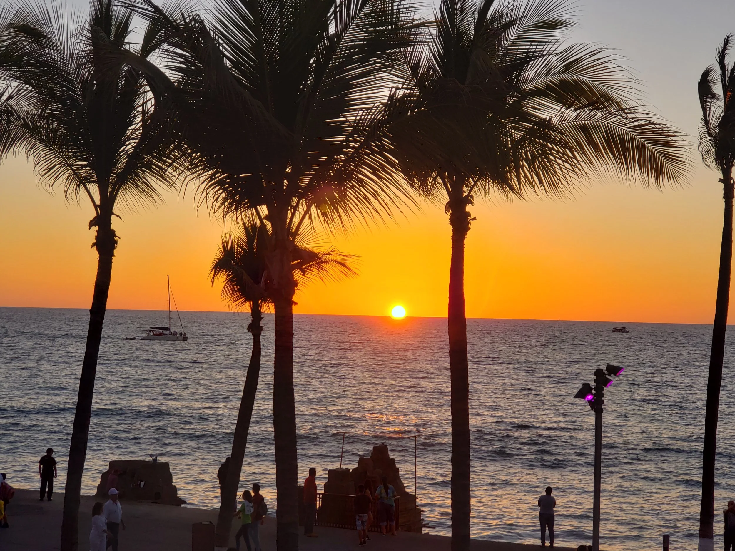 Sunset over the ocean with palm trees in the foreground and boats on the water, people walking along the shore.