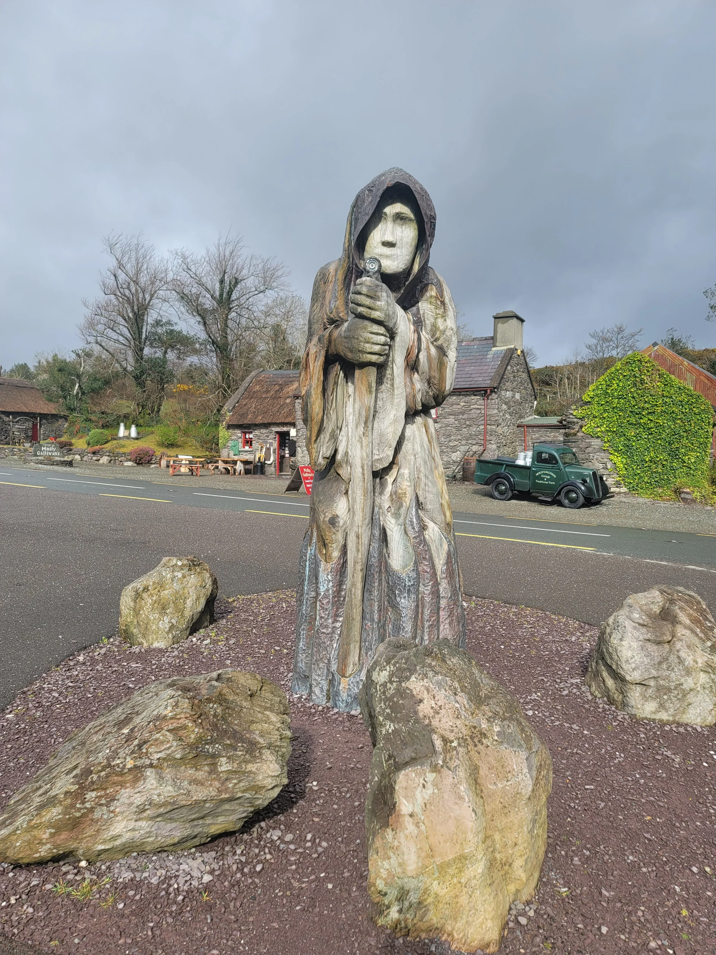 Wooden sculpture of a cloaked figure with a face holding a staff, surrounded by large rocks, positioned on a gravel patch near a roadside with a historic village in the background.