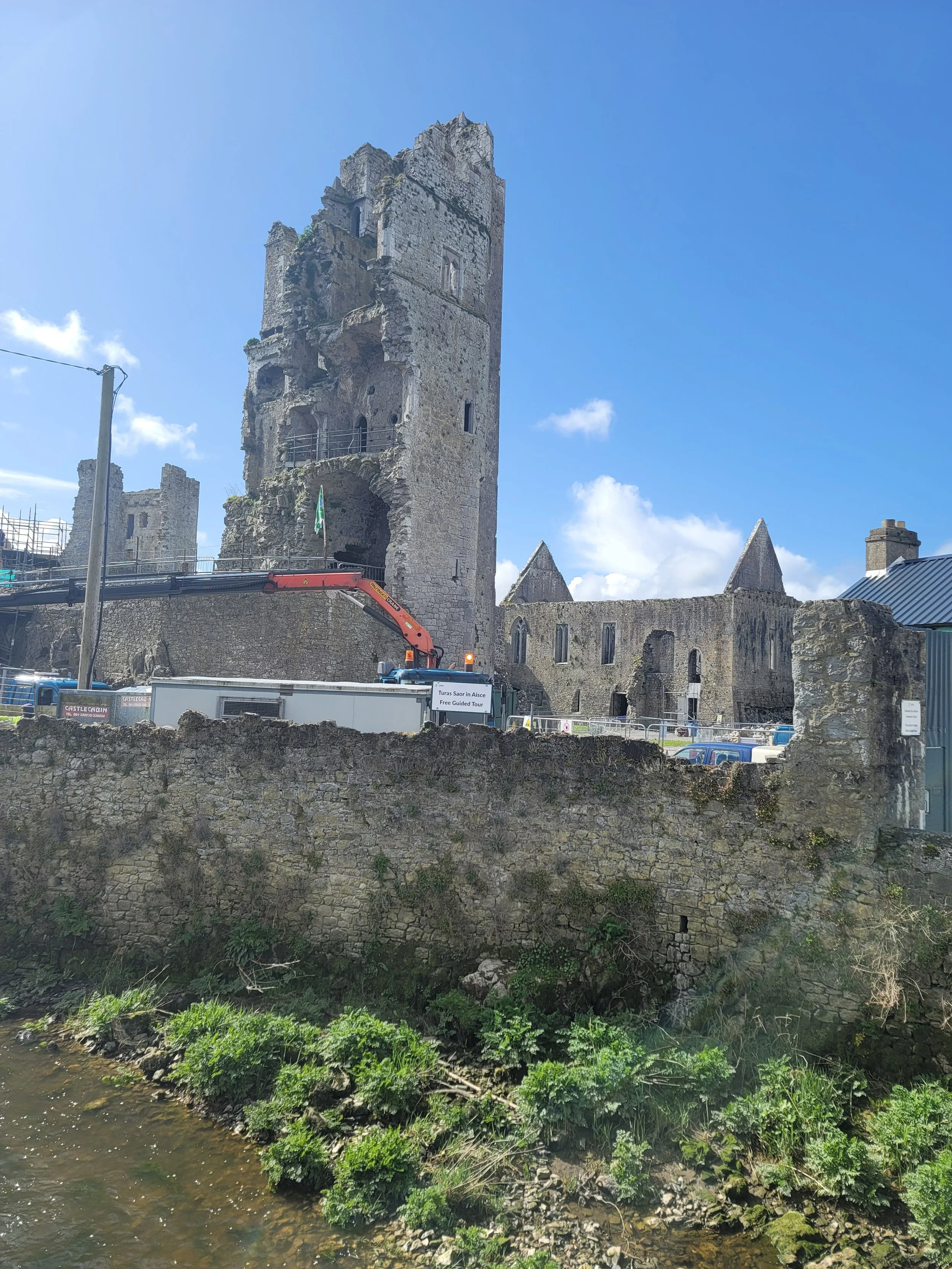 Ancient stone castle ruins with a tall tower, situated near a small river under a bright blue sky with some clouds.