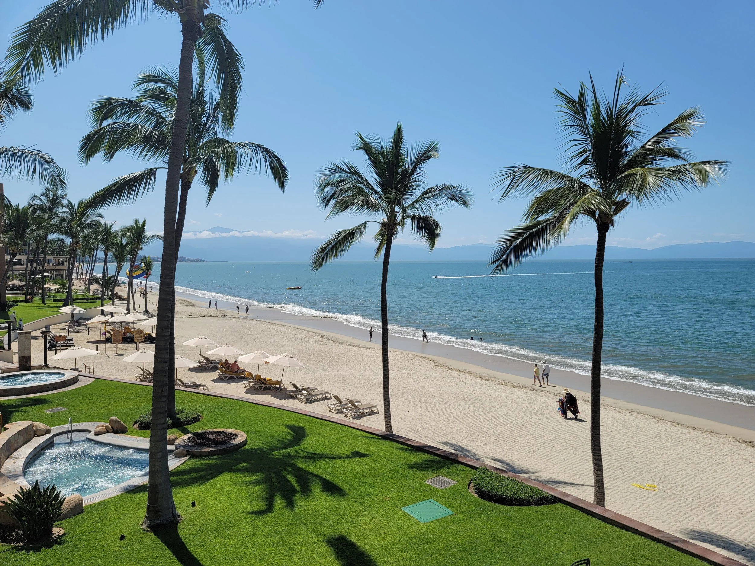 A tropical beach scene with palm trees, sandy shoreline, ocean water, and people walking or lounging under umbrellas, with mountains in the distance.