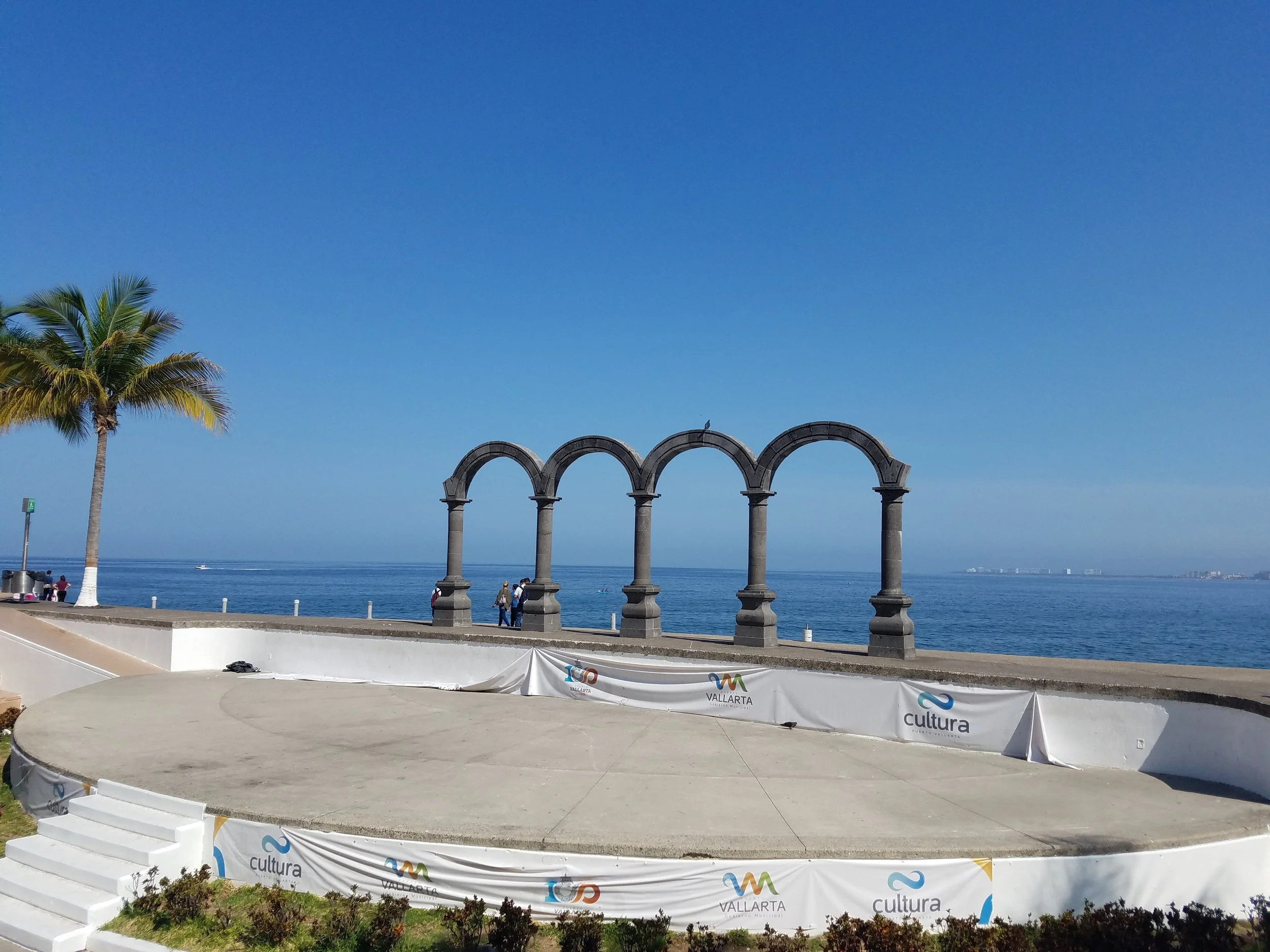 A seaside promenade in Vallarta with a palm tree on the left, a stage with white banners featuring the Burgos Vallarta and Cultura logos, and four stone arches against the ocean and blue sky in the background.