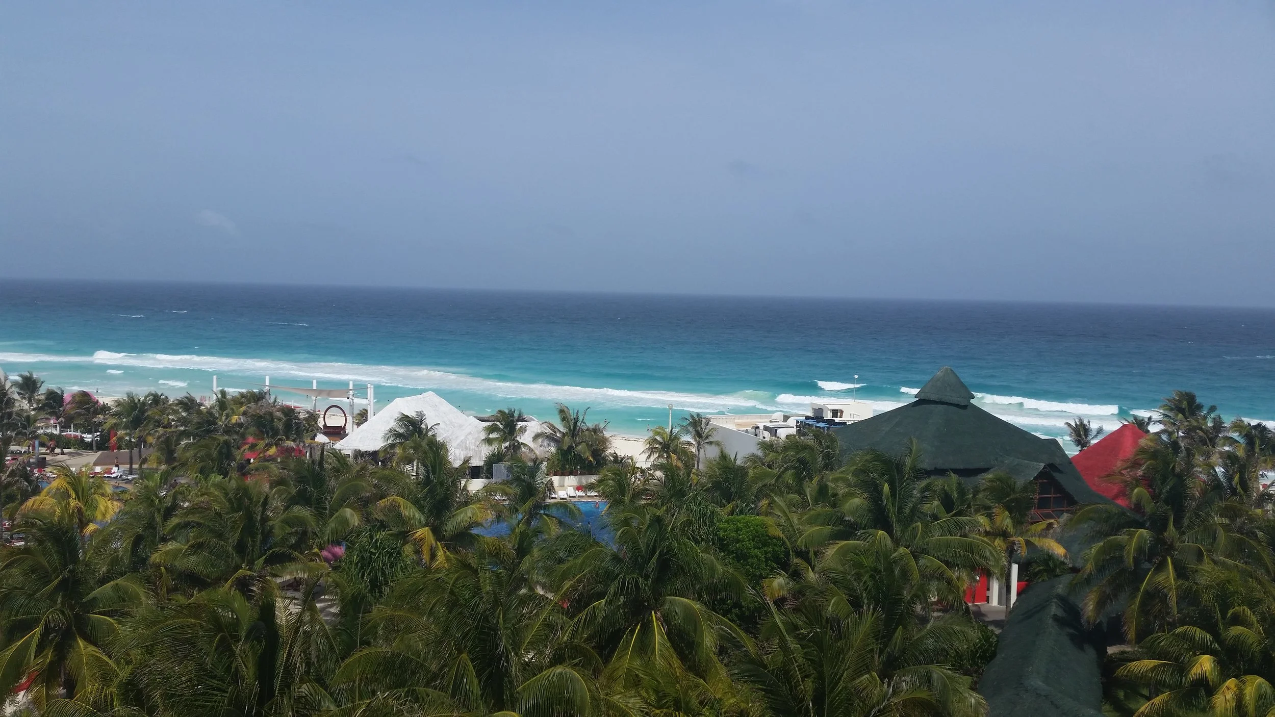 Tropical beach view with blue ocean, waves, palm trees, and resort structures with thatched roofs