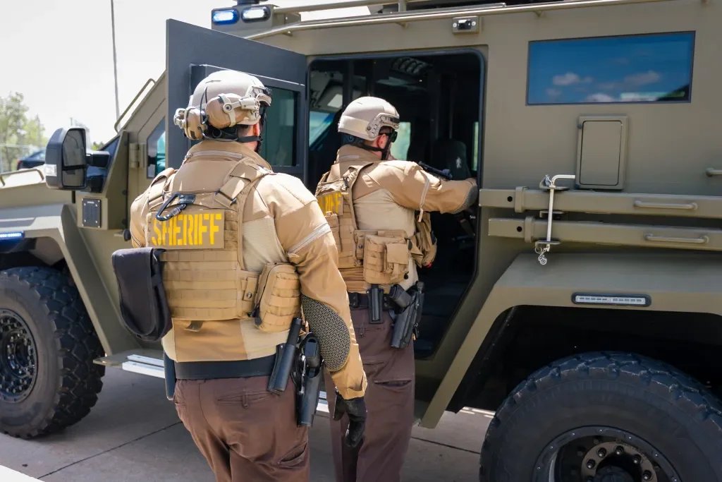 Two sheriff deputies in tactical gear approach a military-style armored vehicle, with one opening the door and the other standing nearby.