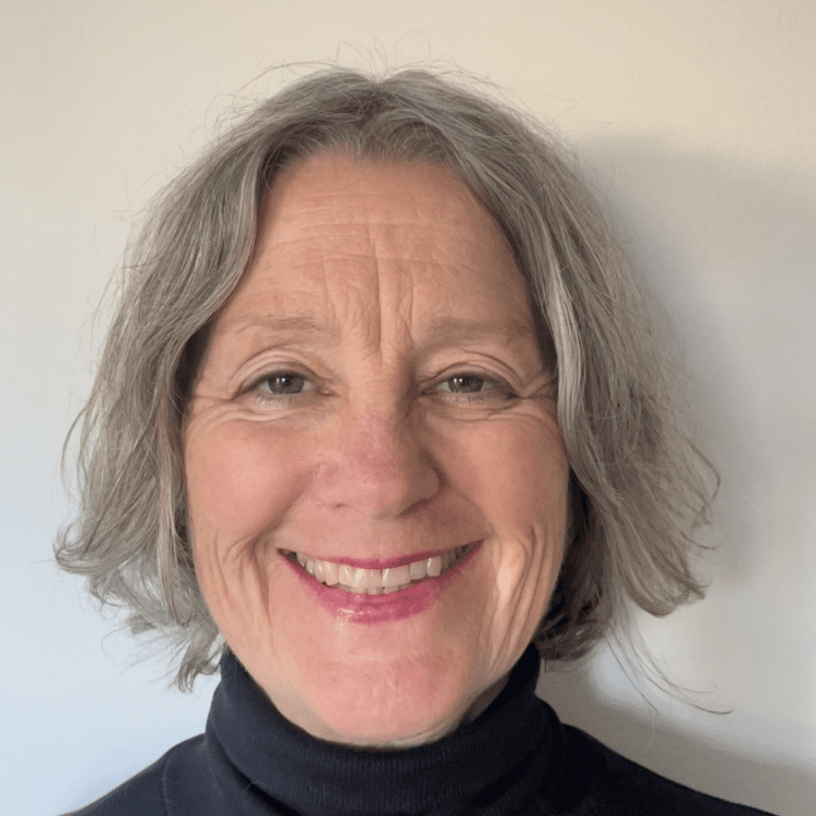 A smiling middle-aged woman with gray, wavy hair wearing a black turtleneck against a plain wall