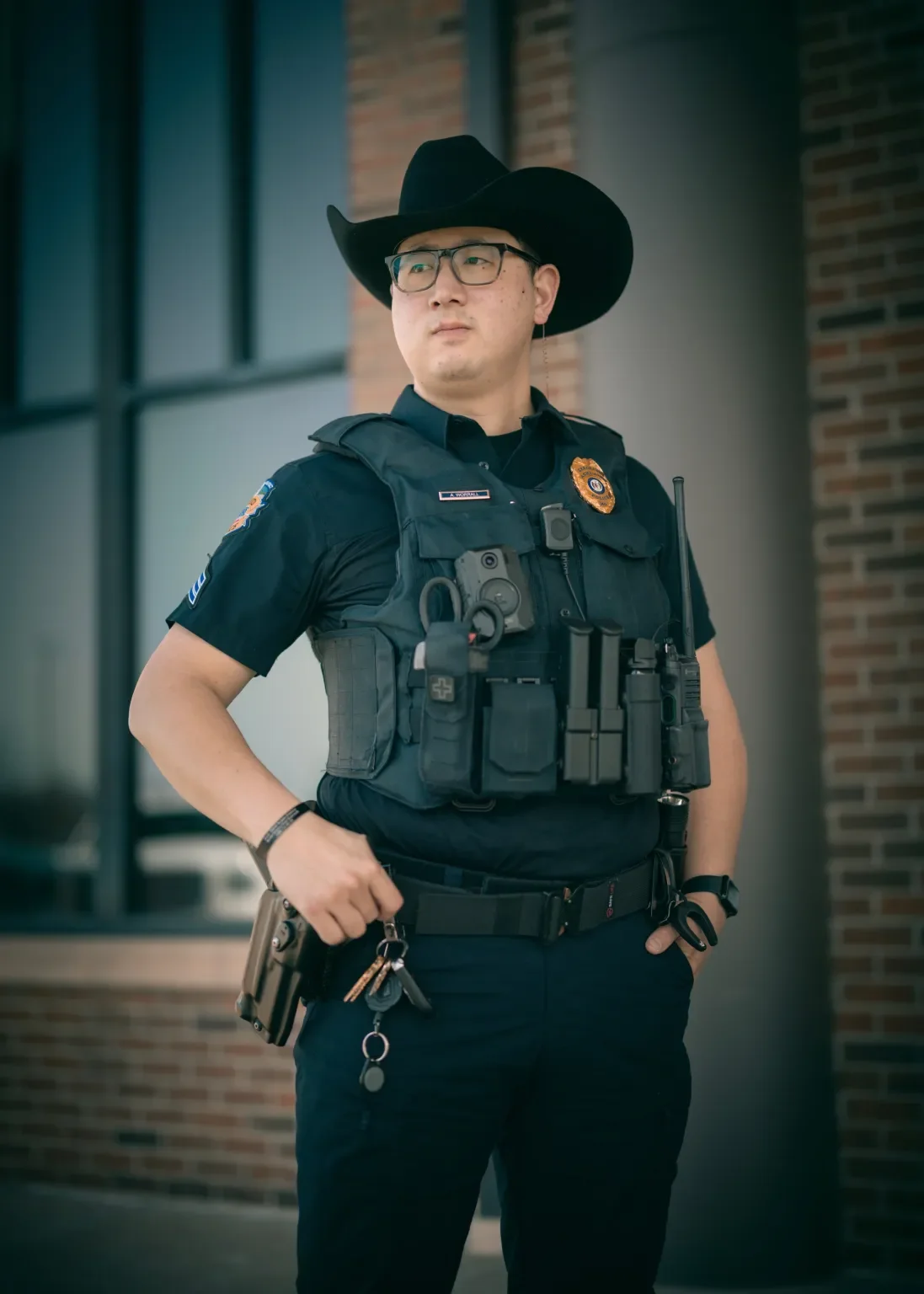 A police officer wearing a cowboy hat, glasses, a black uniform, and a vest with equipment stands with one hand in his pocket outside a building with brick walls and windows.