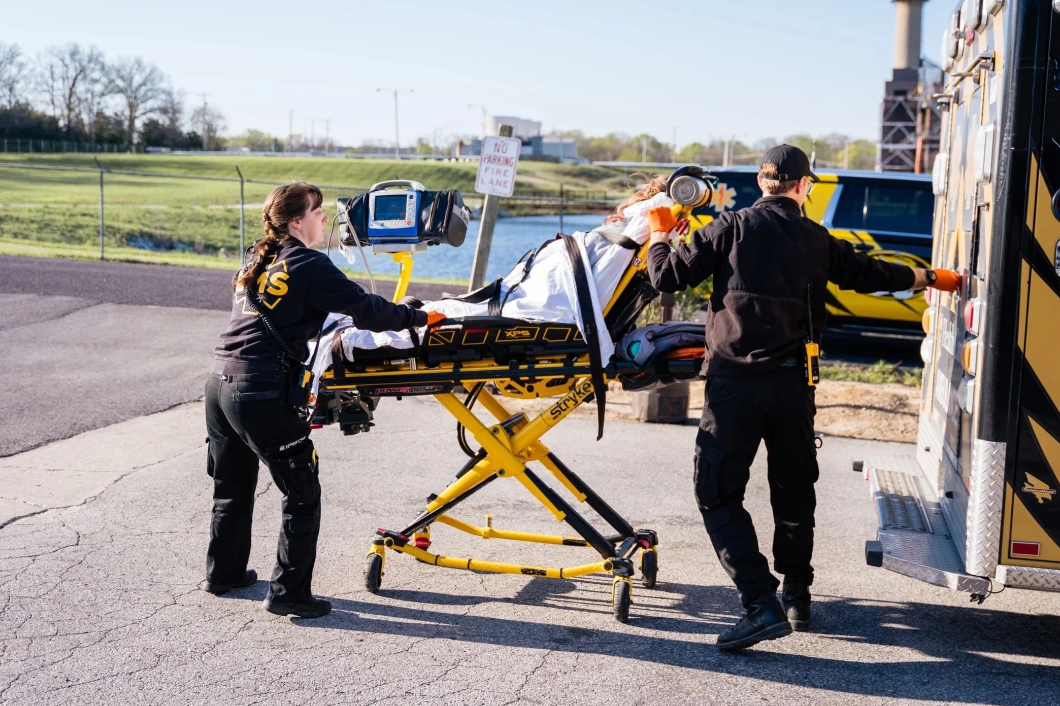 Emergency medical personnel transporting a patient on a stretcher to an ambulance on a paved outdoor area.