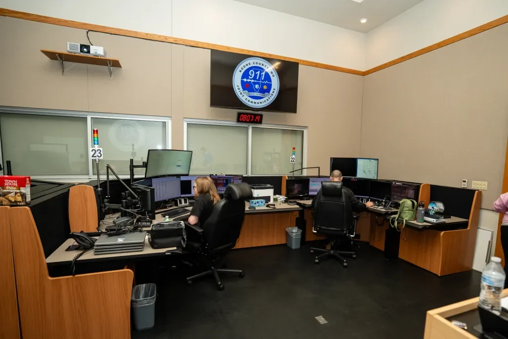 A police emergency communication room with multiple workstations, monitors, and radios. There are two people working at their desks, with the Boone County Sheriff's Office logo on a screen above.