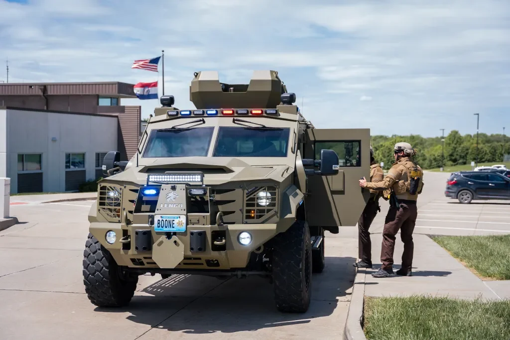 Military police officers near an armored vehicle in a parking lot, with American and Missouri flags in the background.
