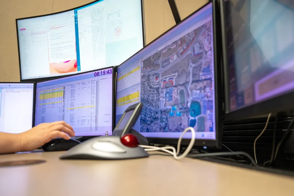 Multiple computer monitors displaying spreadsheets, maps, and email clients in a control room setting.