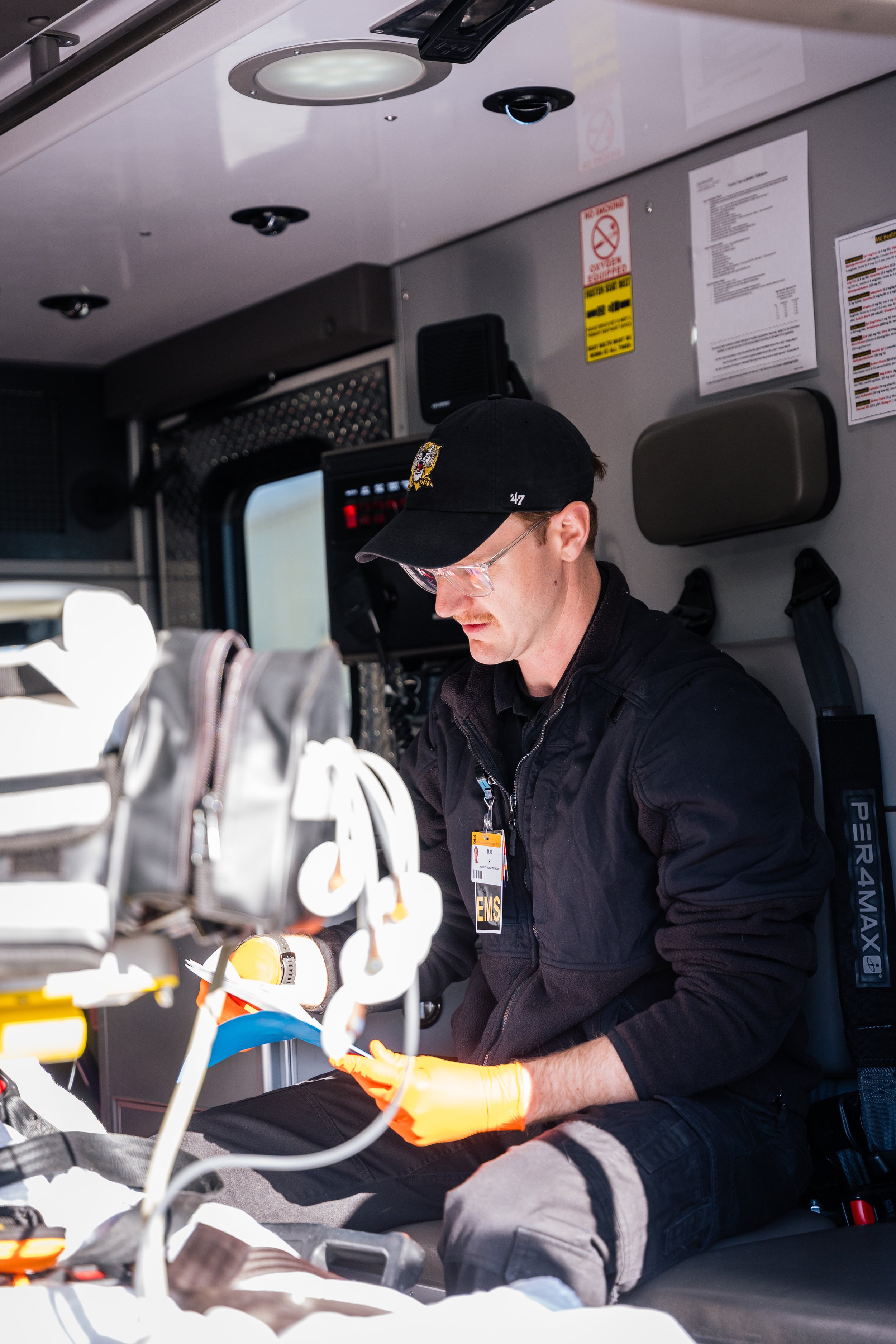 An emergency medical technician (EMT) sitting in the back of an ambulance, wearing orange gloves and a black cap, working with medical equipment.