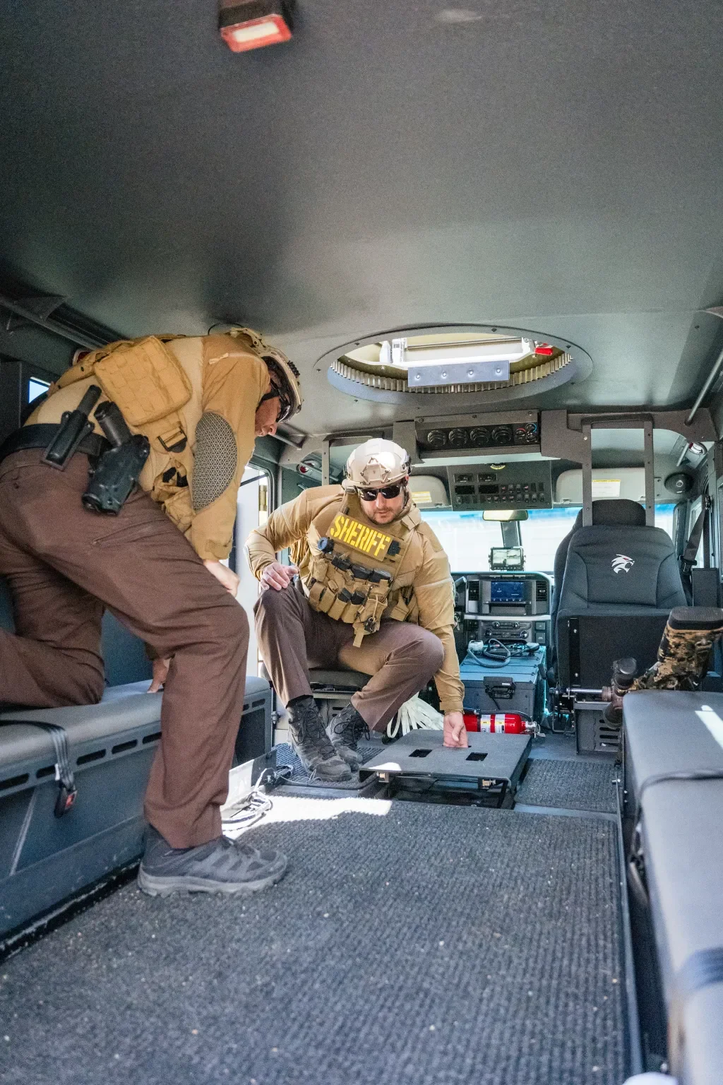 Two law enforcement officers in tactical gear inside a police or emergency vehicle, preparing or inspecting equipment.
