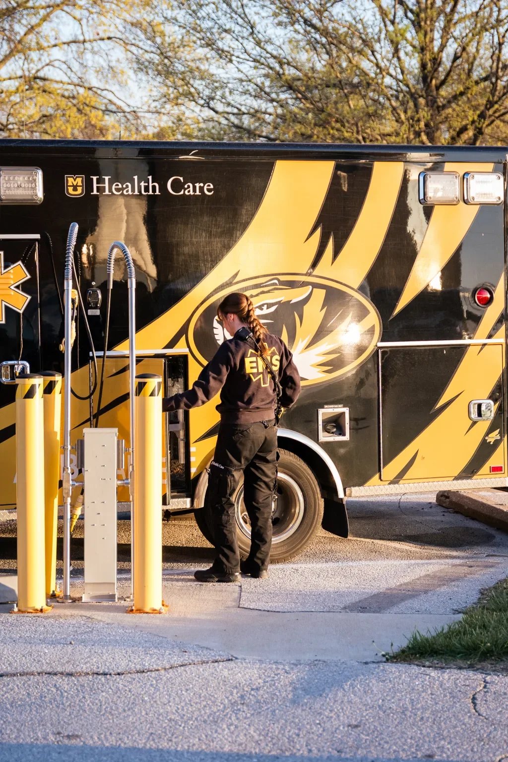 A female emergency medical technician (EMT) in a black uniform with 'EMS' written on the back, standing outside a black emergency vehicle with gold stripes and a tiger logo, connected to medical equipment at a dialysis or on-site health care clinic s