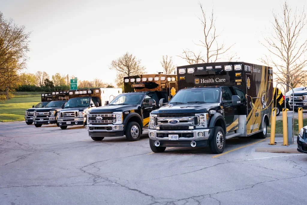 A lineup of emergency medical vehicles, including ambulances and support trucks, parked in a lot with trees in the background.